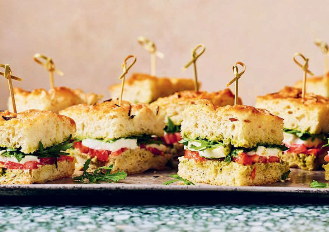 Sandwiches with tomato, lettuce, and cheese on focaccia bread, secured with toothpicks, on a serving platter.