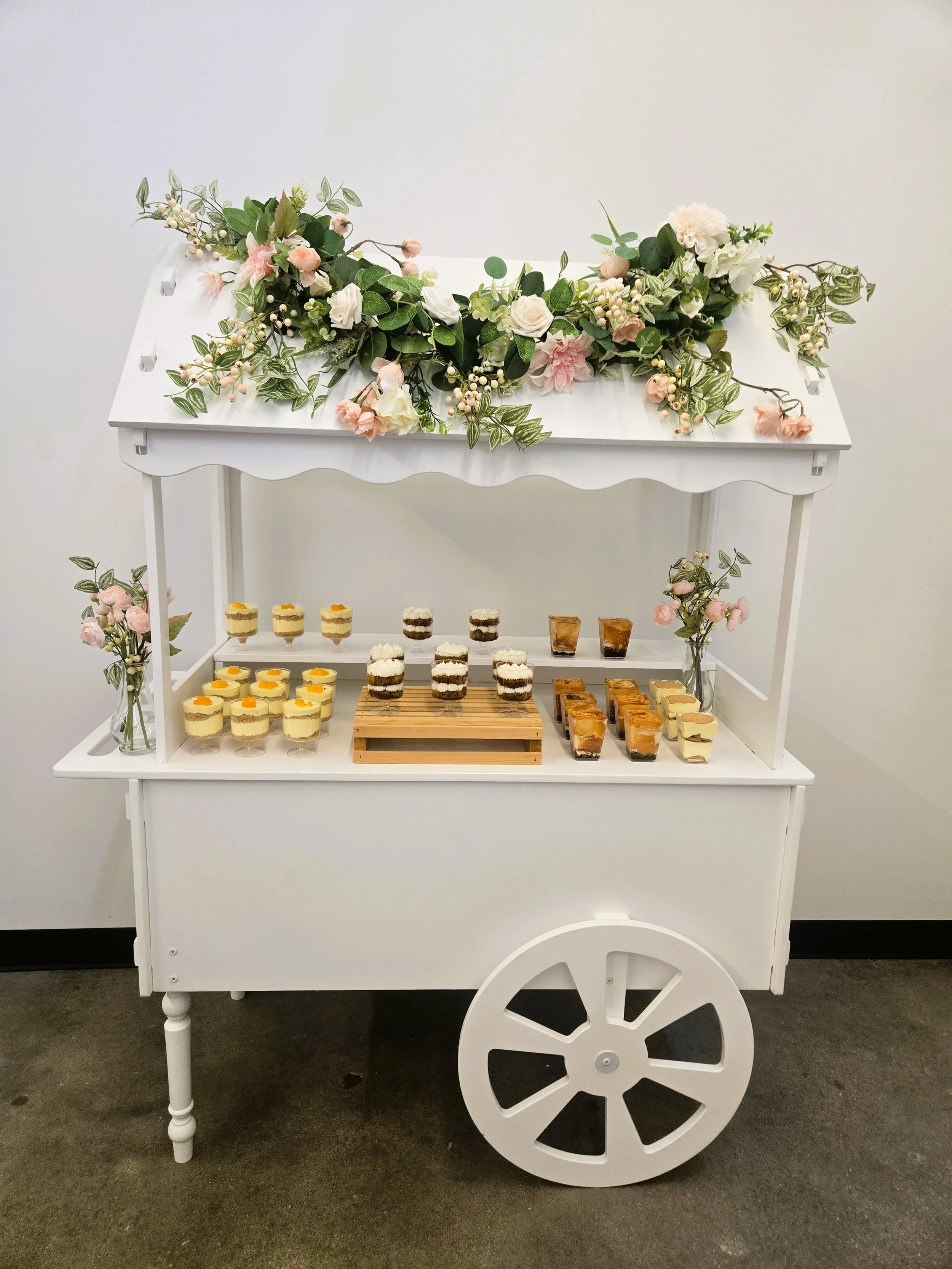 White dessert cart decorated with pink and white flowers and green foliage, displaying various mini desserts such as cheesecakes, layered parfaits, and parfaits on the shelves.