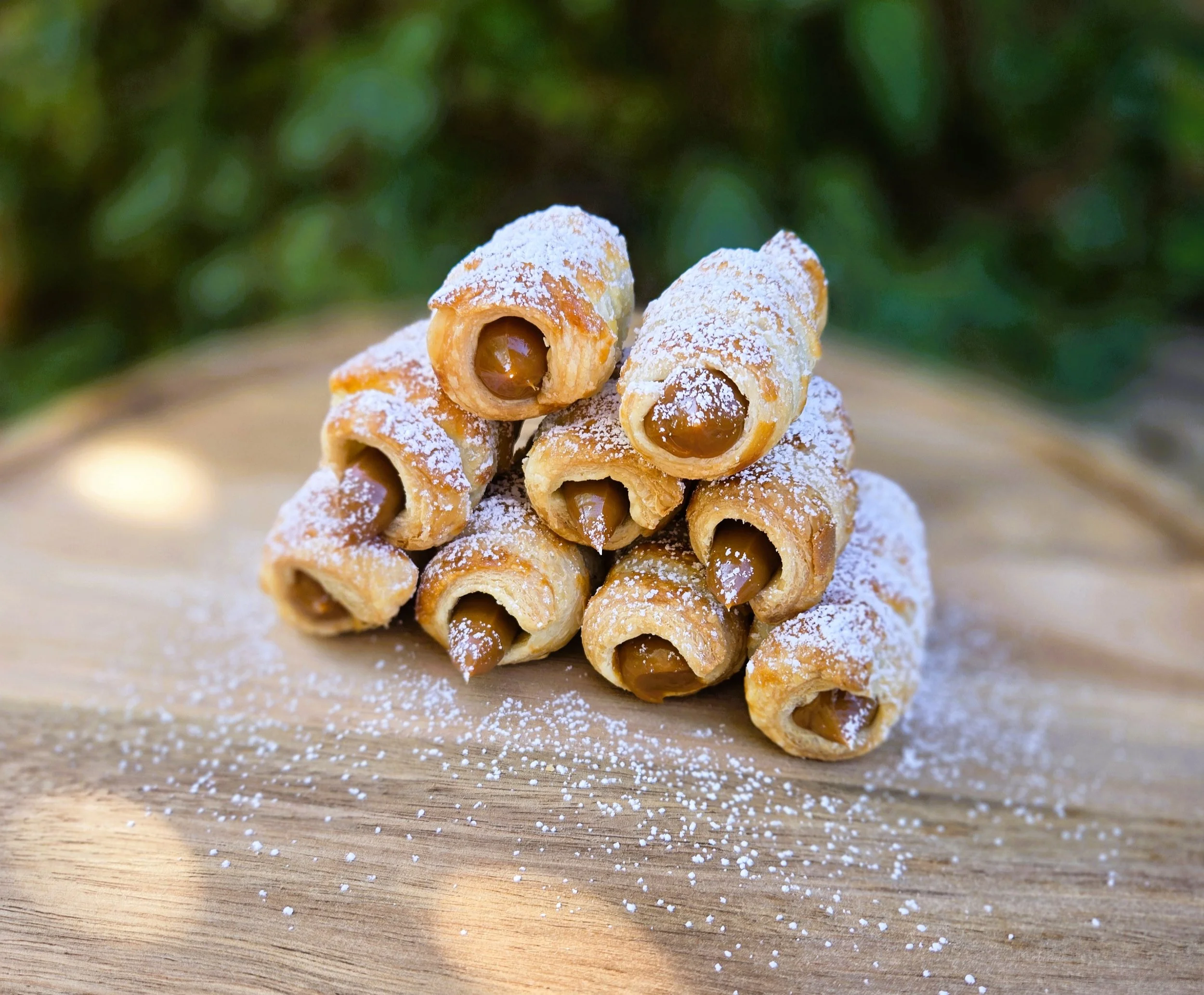 Stack of cannoli with powdered sugar on a wooden surface, outdoors.