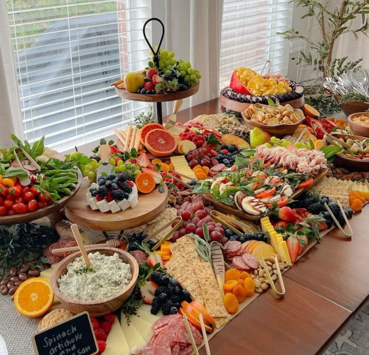 A large spread of various fresh fruits, vegetables, cheeses, meats, and dips on a wooden table near a window.
