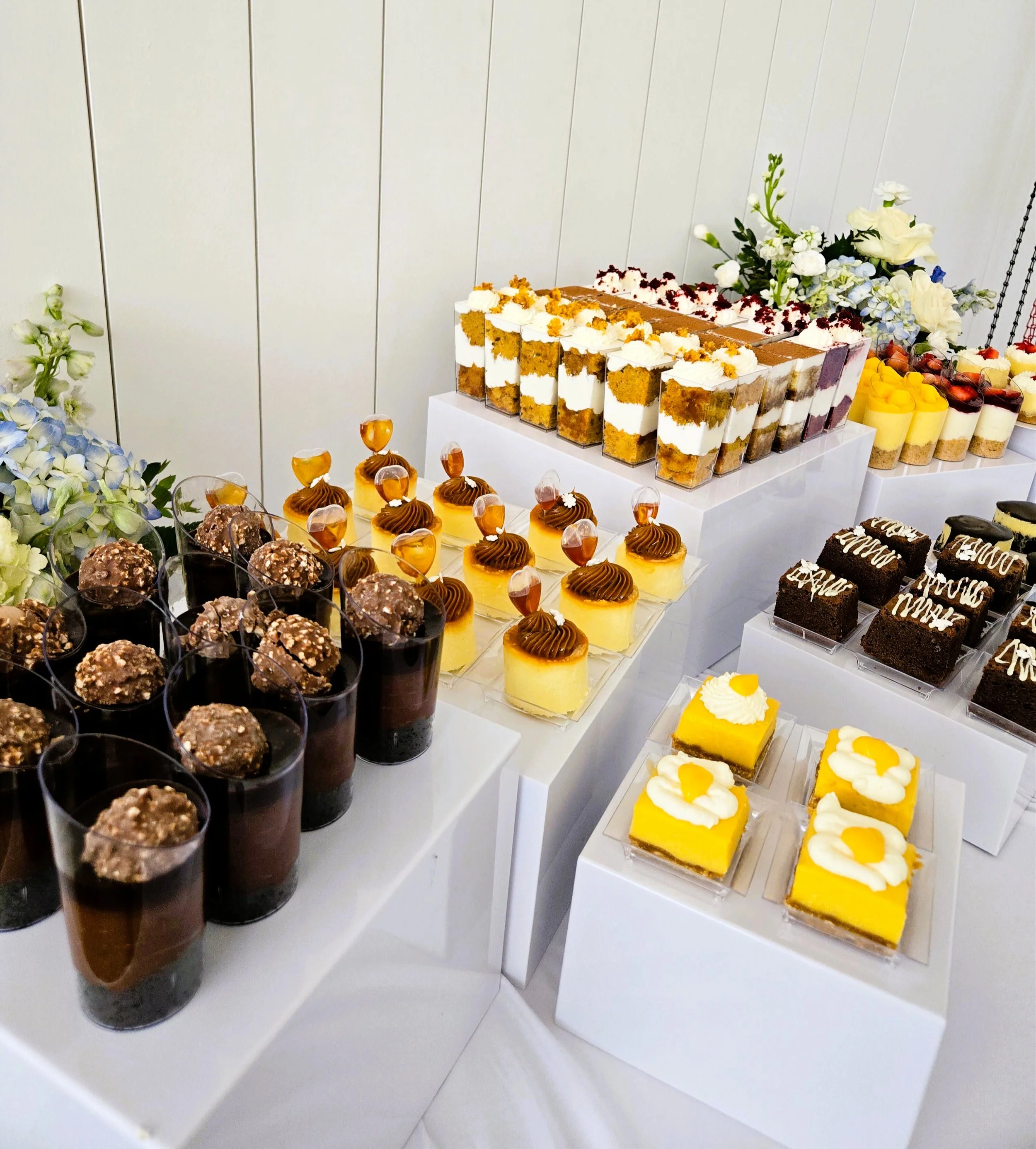 Assorted desserts including chocolate mousse cups, tiramisu, lemon bars, and small cakes, decorated with flowers at a catering event.