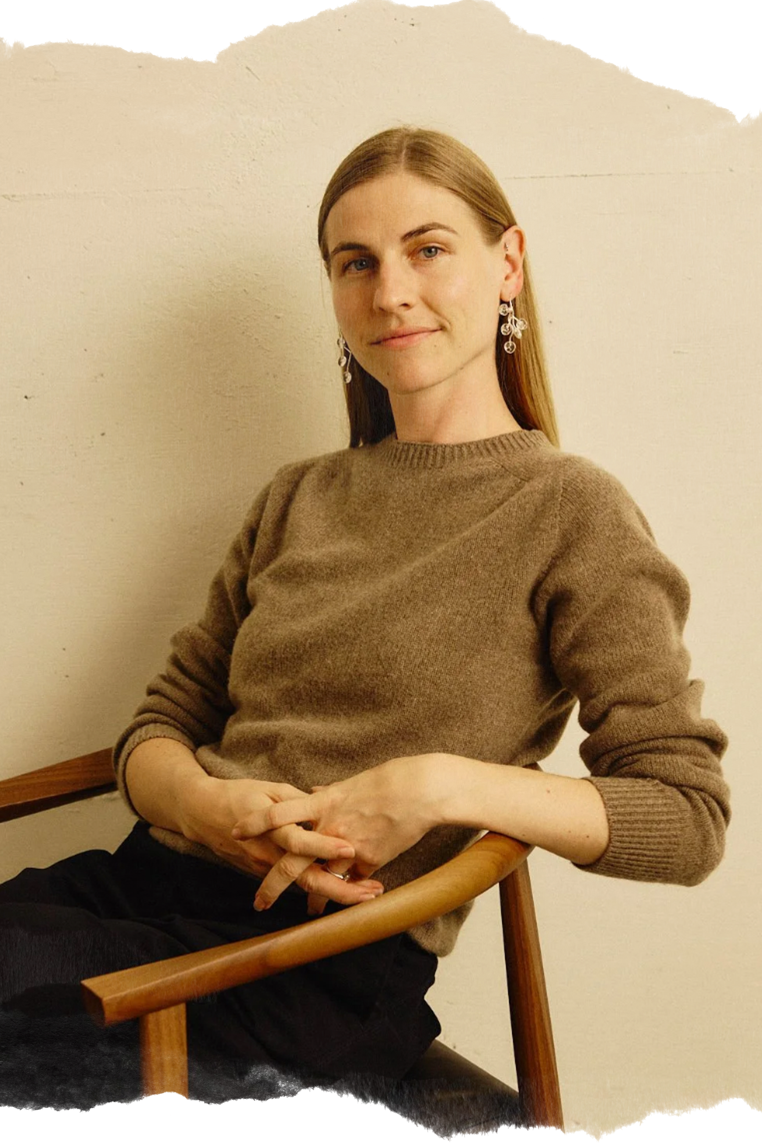 A woman with long hair wearing earrings and a brown sweater, sitting on a wooden chair with a neutral expression and hands resting on her lap, in front of a plain light-colored wall.
