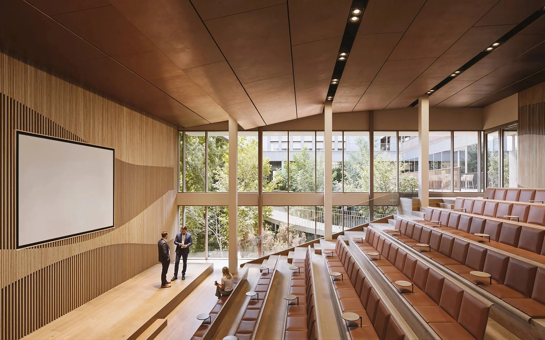 Modern auditorium with tiered seating, a large blank screen, and floor-to-ceiling windows overlooking trees.