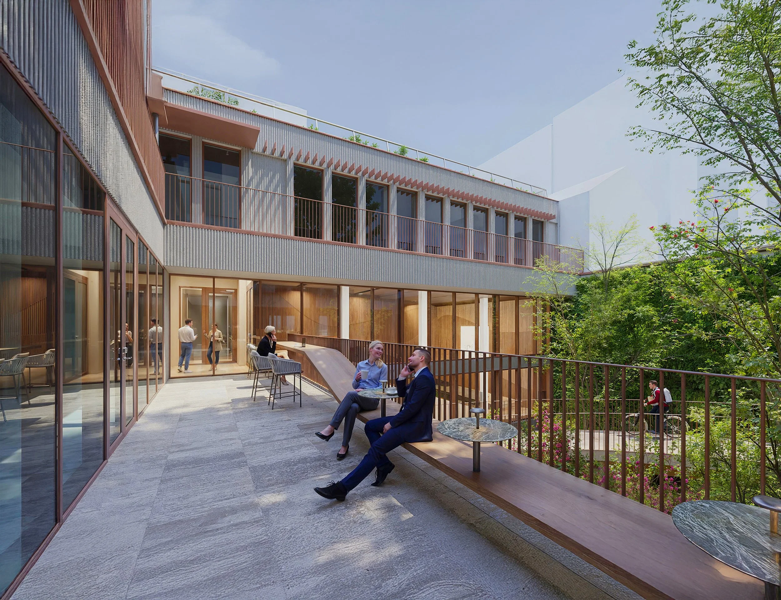 Modern outdoor terrace with people sitting and chatting, surrounded by glass walls and lush green trees, near a multi-story building with large windows.
