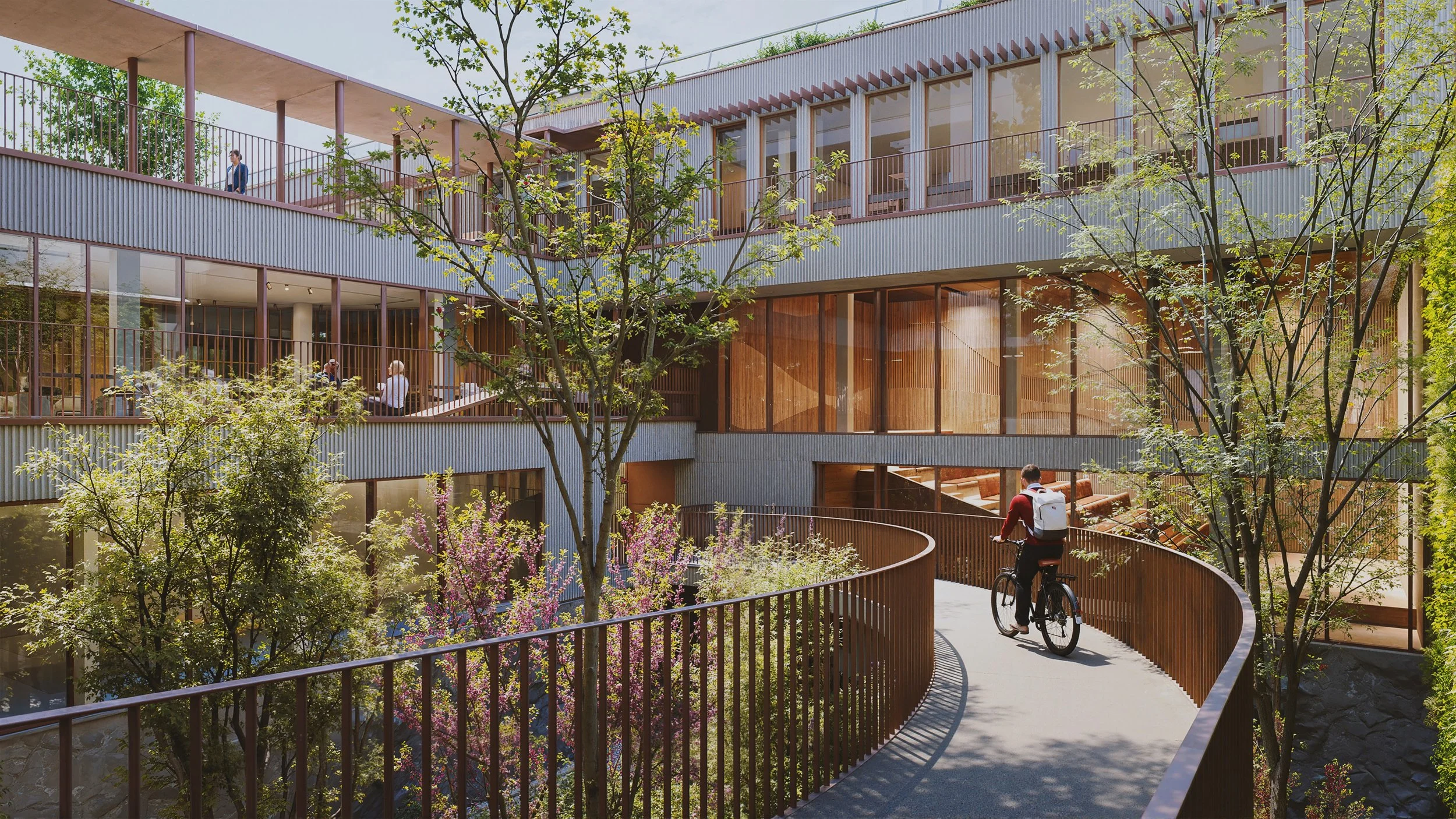 A person riding a bicycle on a winding pathway surrounded by trees and modern multi-story buildings with glass windows and wooden accents.
