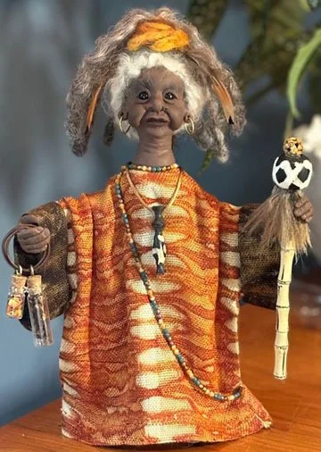 A doll dressed in traditional, colorful clothing with long, curly, gray hair, holding a small staff with a carved skull on top.