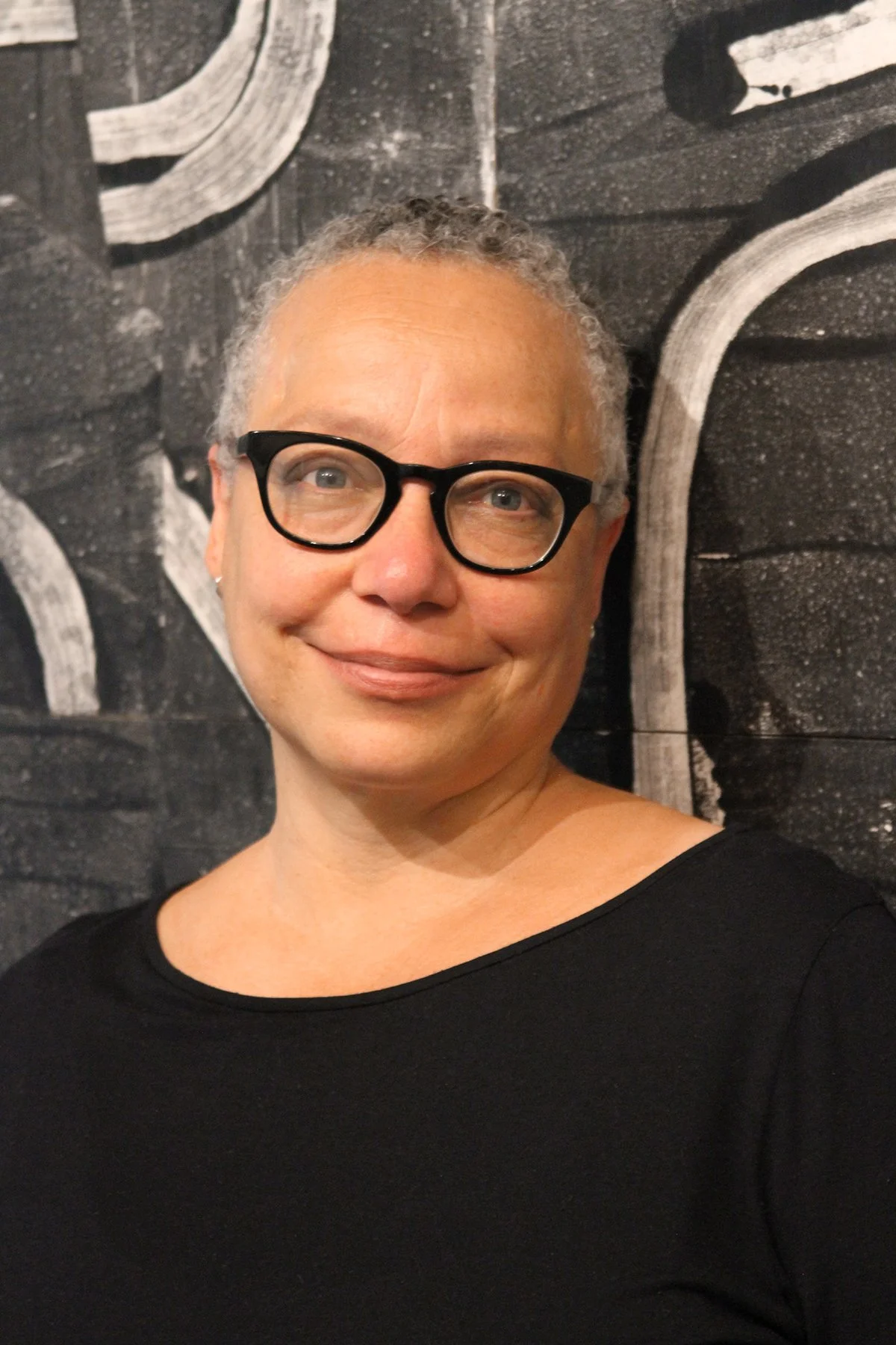 A woman with short gray hair, wearing black glasses and a black top, smiling in front of a textured black and white abstract wall.