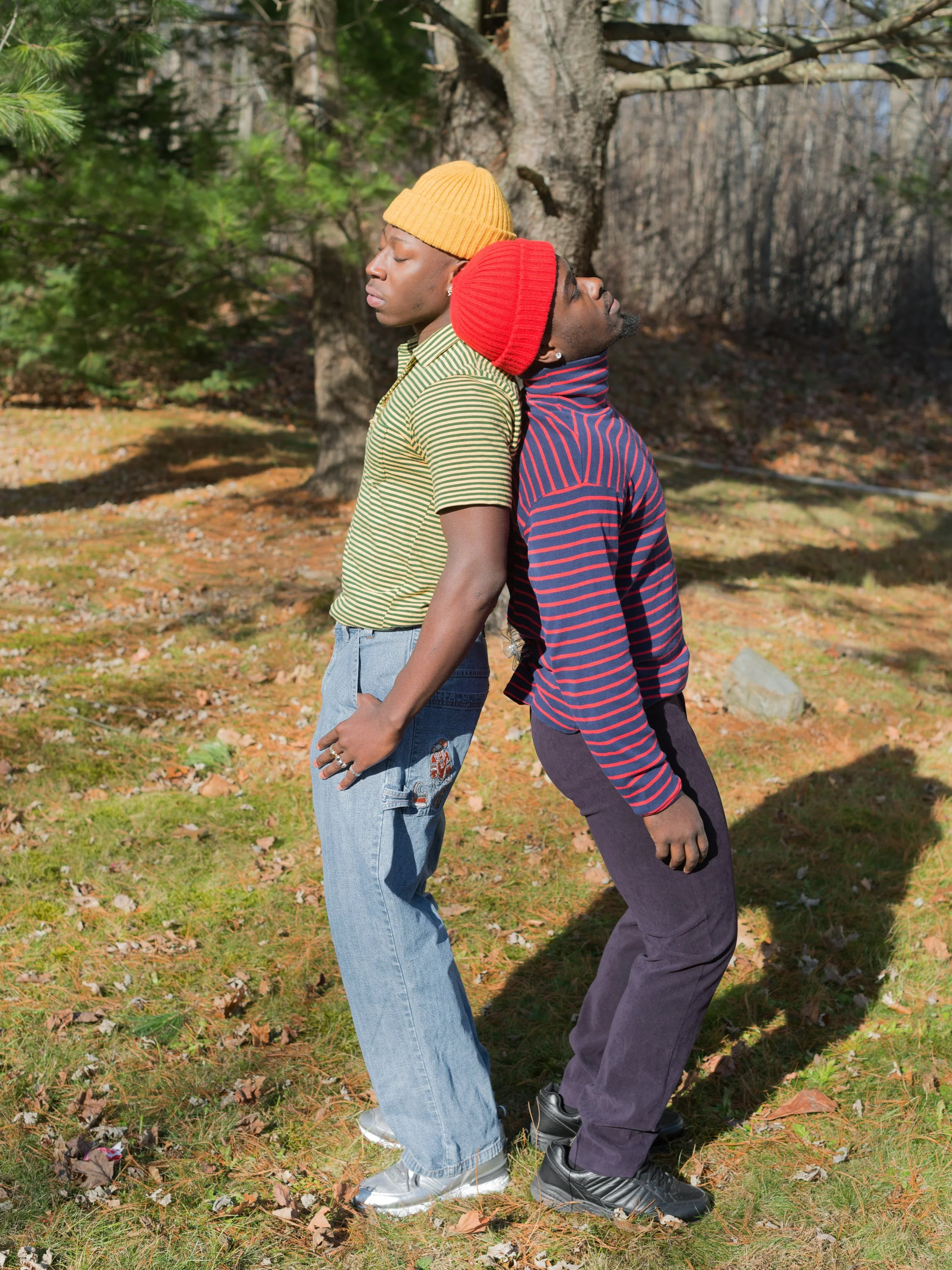Two men stand back-to-back outdoors in a park on a sunny day, leaning against each other with their eyes closed. One wears a yellow beanie and a green striped shirt, the other a red beanie and a blue striped shirt.