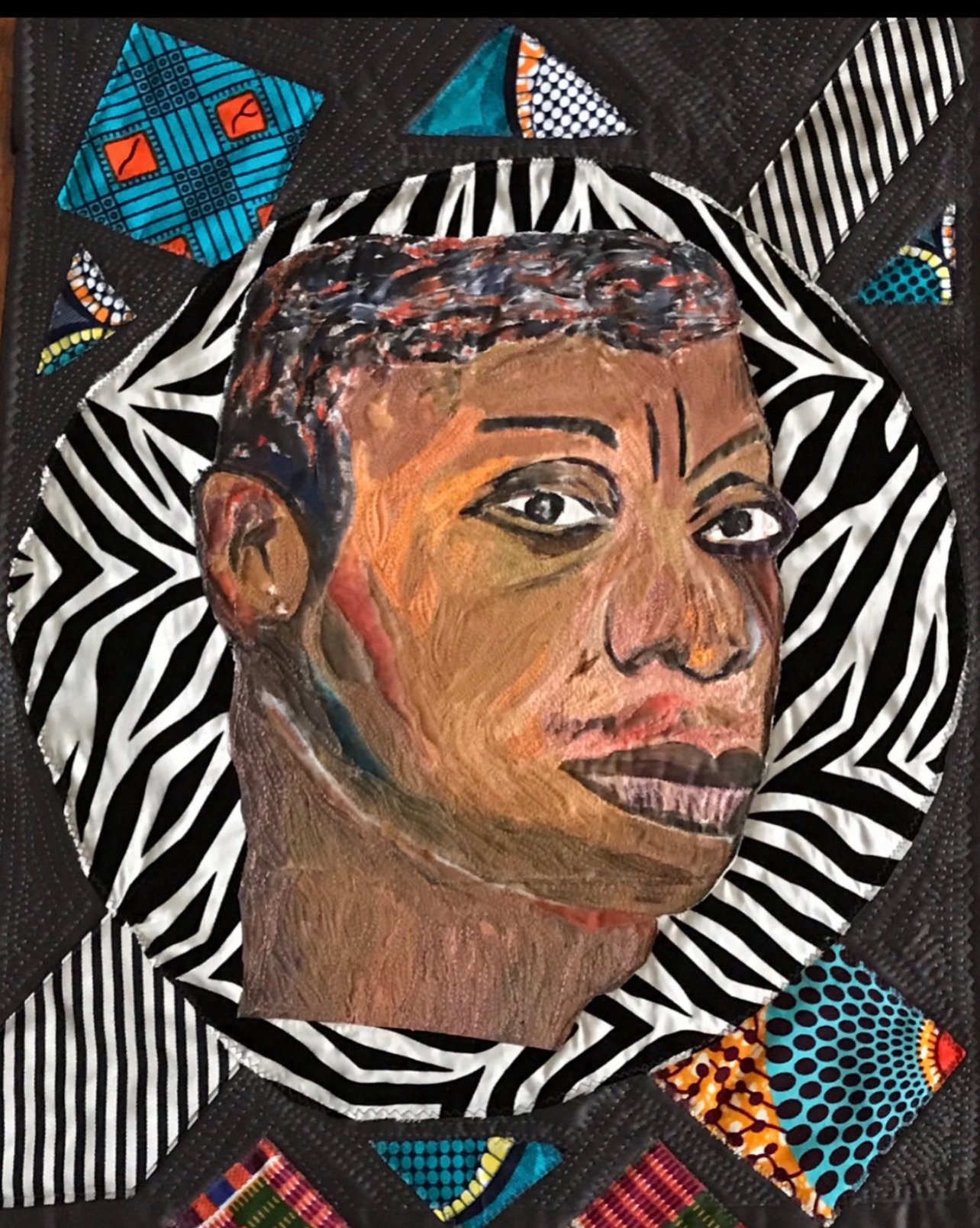 A mixed media artwork of a woman's face with dark hair, almond-shaped eyes, and full lips, surrounded by a vibrant, patterned quilt with geometric and African-inspired designs.
