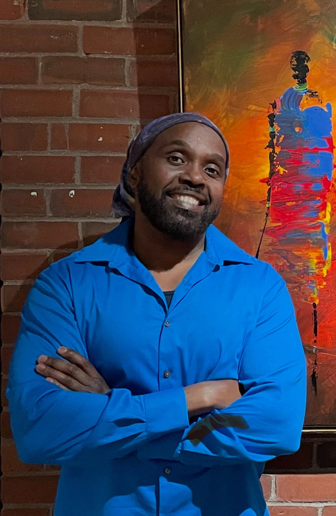 A smiling man with a beard and mustache, wearing a blue dress shirt and a headscarf, standing with arms crossed in front of a colorful abstract painting and brick wall.