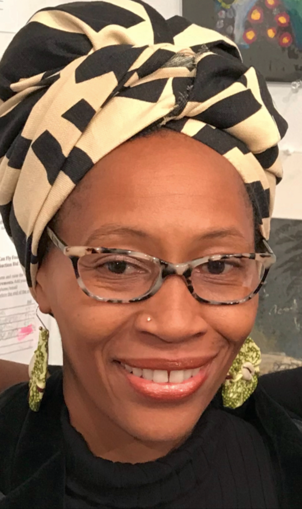 A woman wearing a leopard-print headwrap, glasses, and green earrings, smiling at the camera.