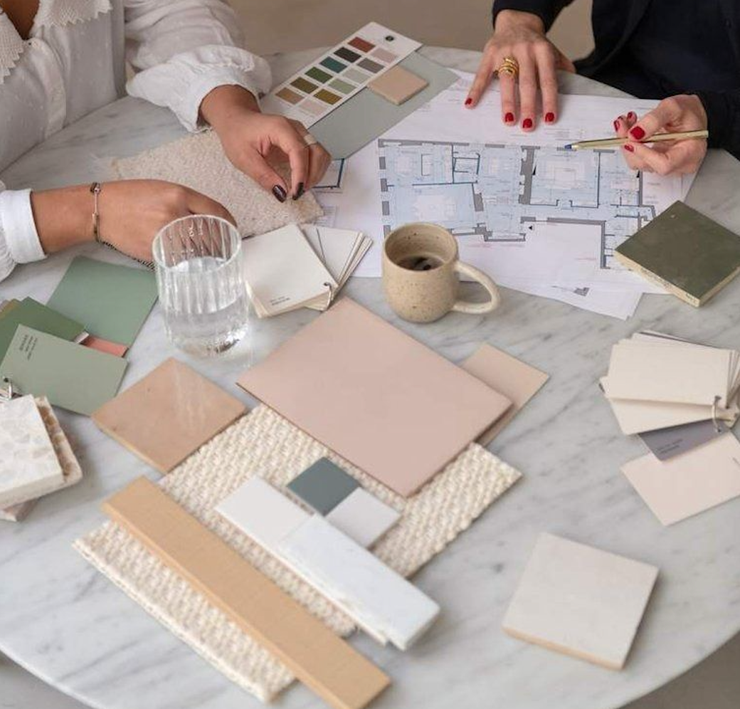 Two people discussing interior design or architecture plans at a marble table, with color swatches, fabric samples, blueprints, and notebooks spread out on the table.