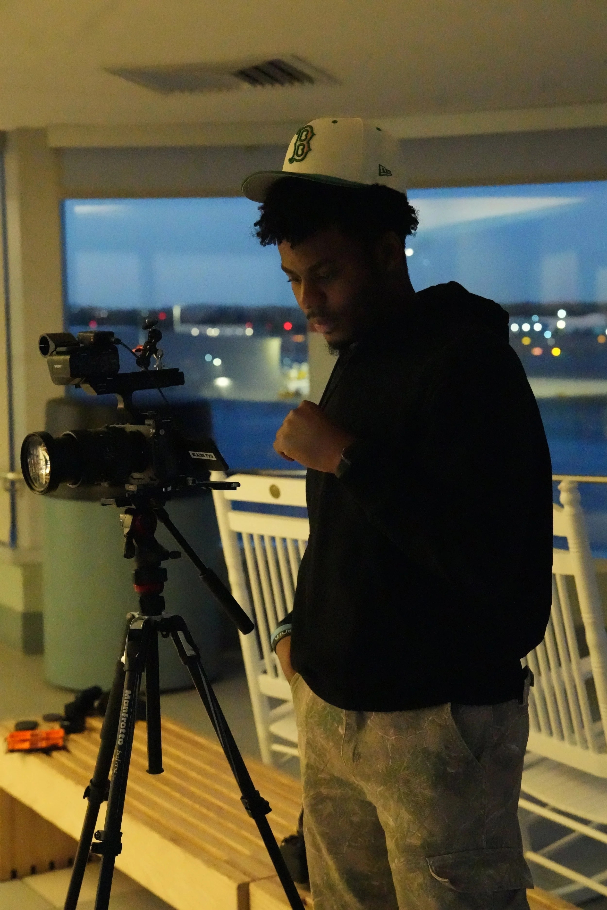 A young man wearing a white cap, black hoodie, and camouflage shorts standing outdoors at dusk, adjusting a professional video camera mounted on a tripod.