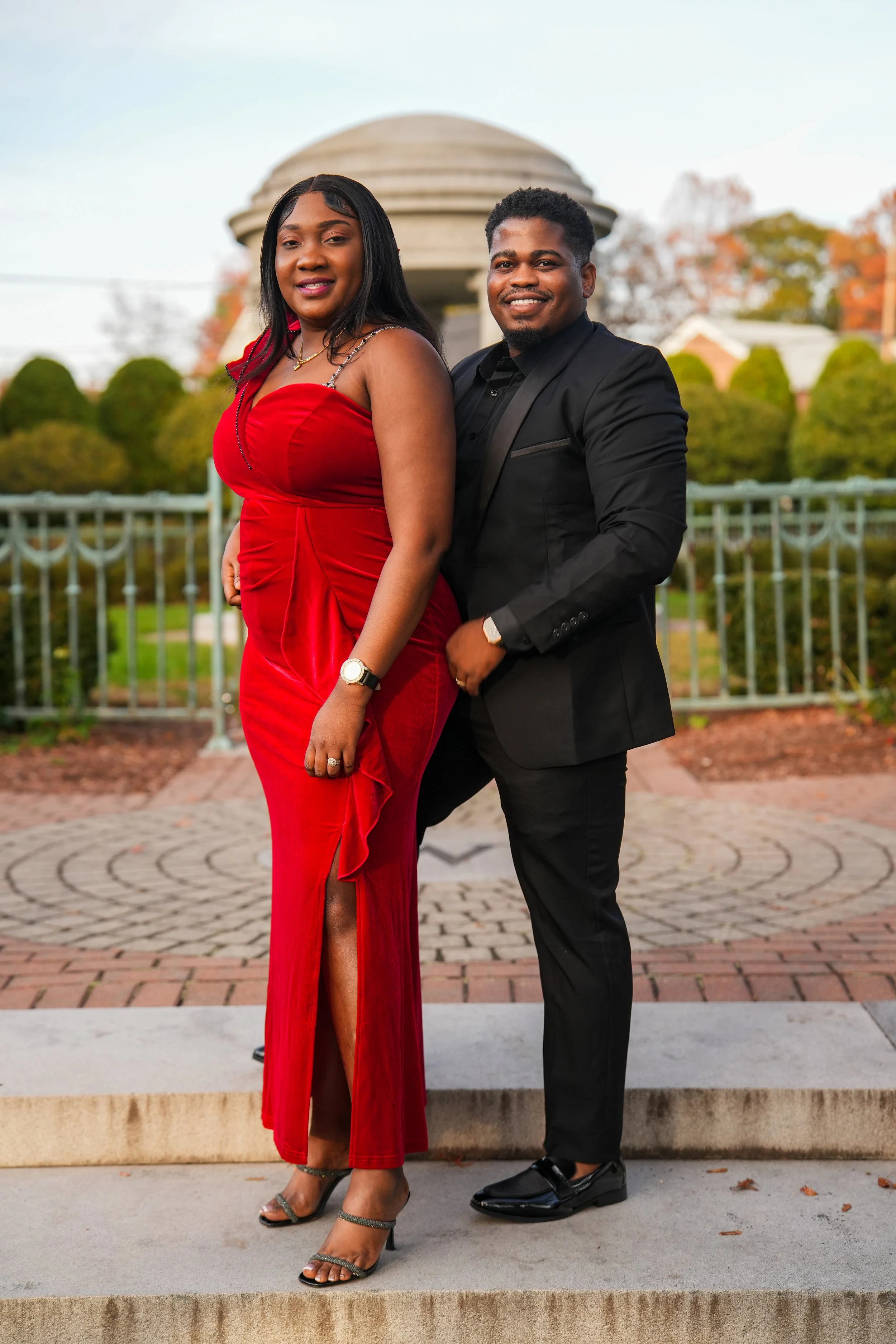 A stylishly dressed couple stands outdoors on a brick walkway with a domed building and autumn trees in the background. The woman wears a red evening gown with a slit, and the man is in a black suit with shiny black shoes anniversary photoshoot inspo
