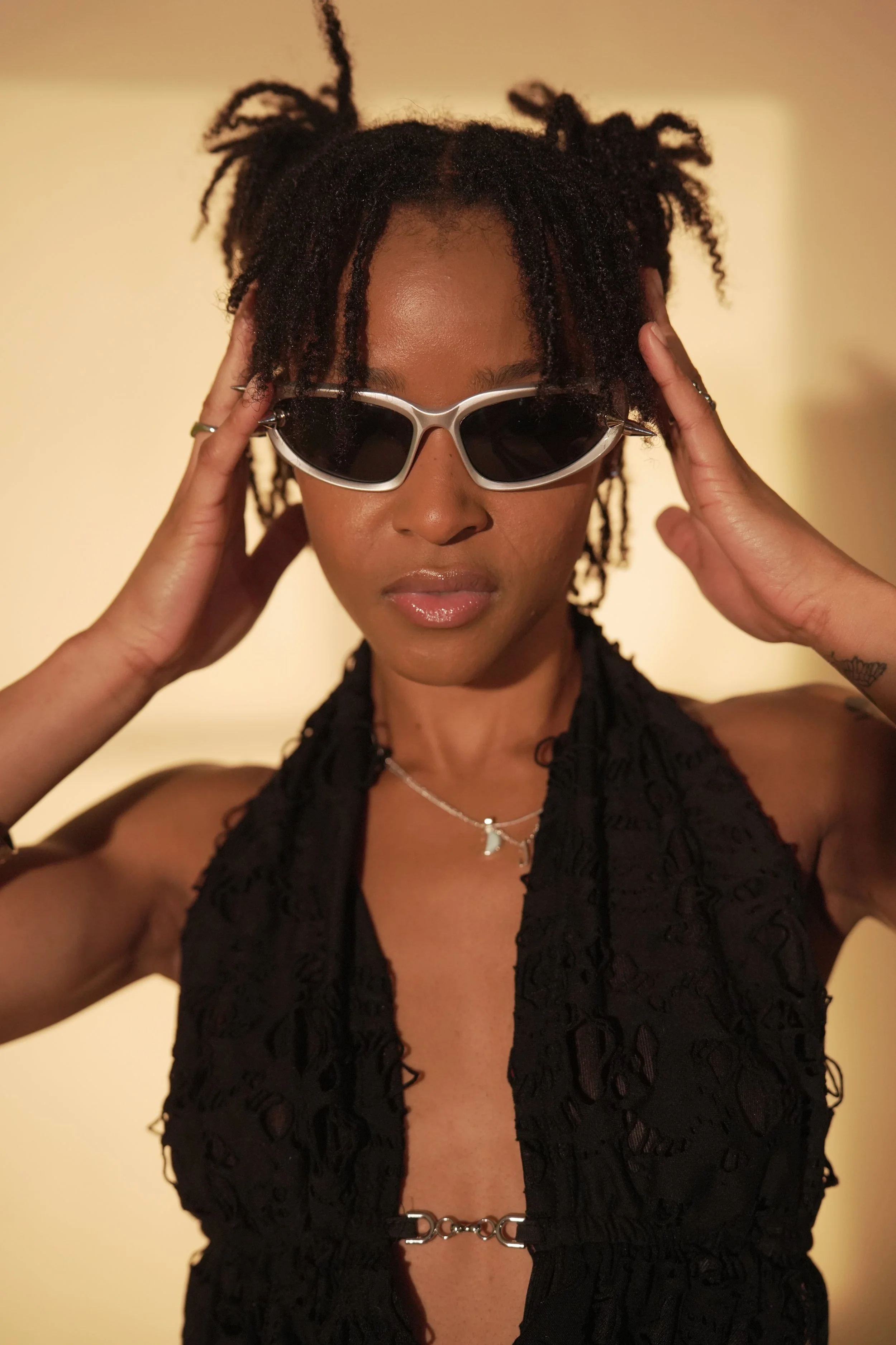 A woman with dark skin wearing large, white sunglasses and a black lace halter top. She holds her hands to the sides of her head, with her eyes covered by sunglasses, in front of a plain, beige background.