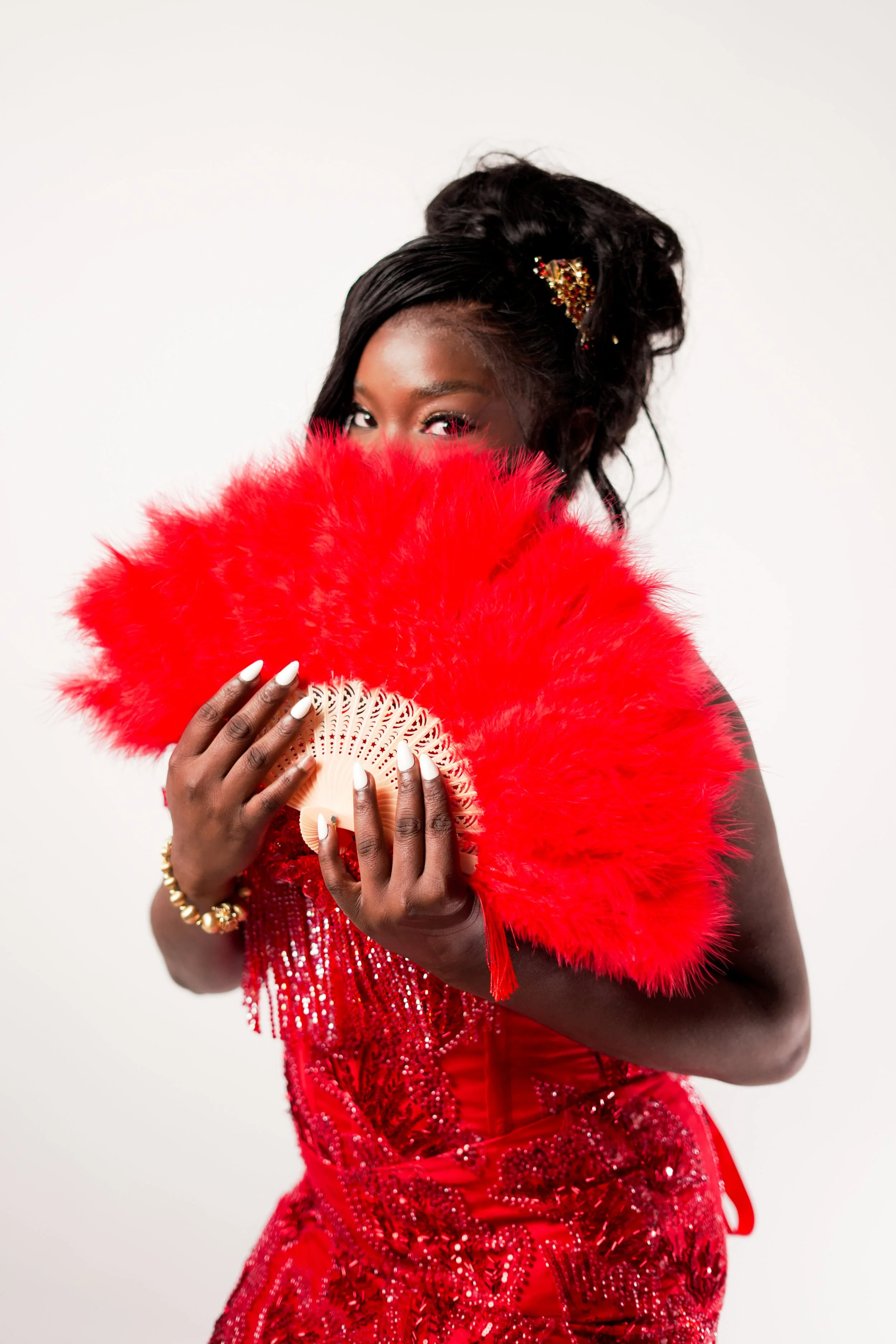 A woman with dark skin wearing a red sequined dress and gold jewelry, partially hiding her face behind a large red feather fan, with her eyes visible and looking at the camera.