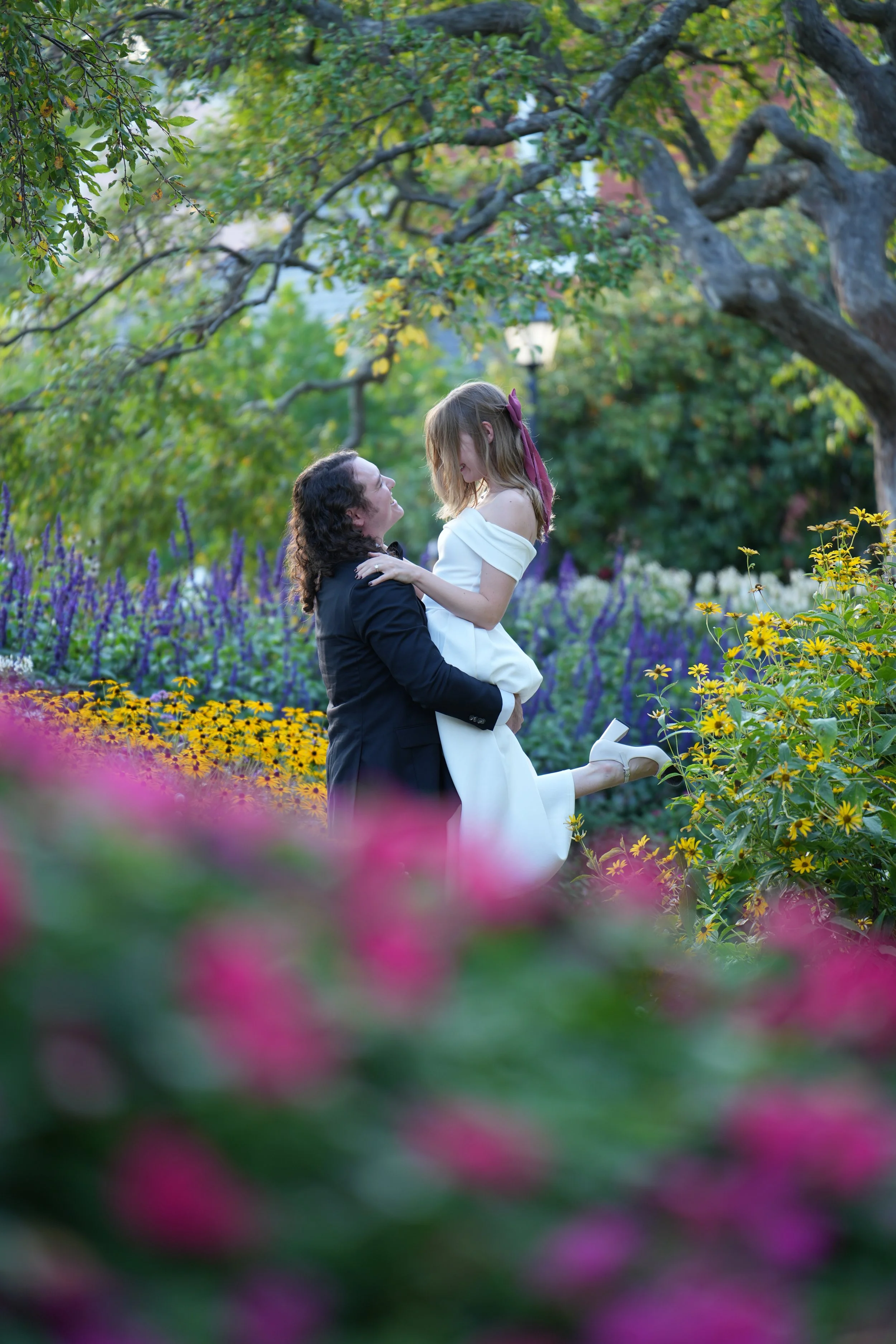 A man lifting a woman in a white dress in a vibrant garden filled with colorful flowers and green trees.