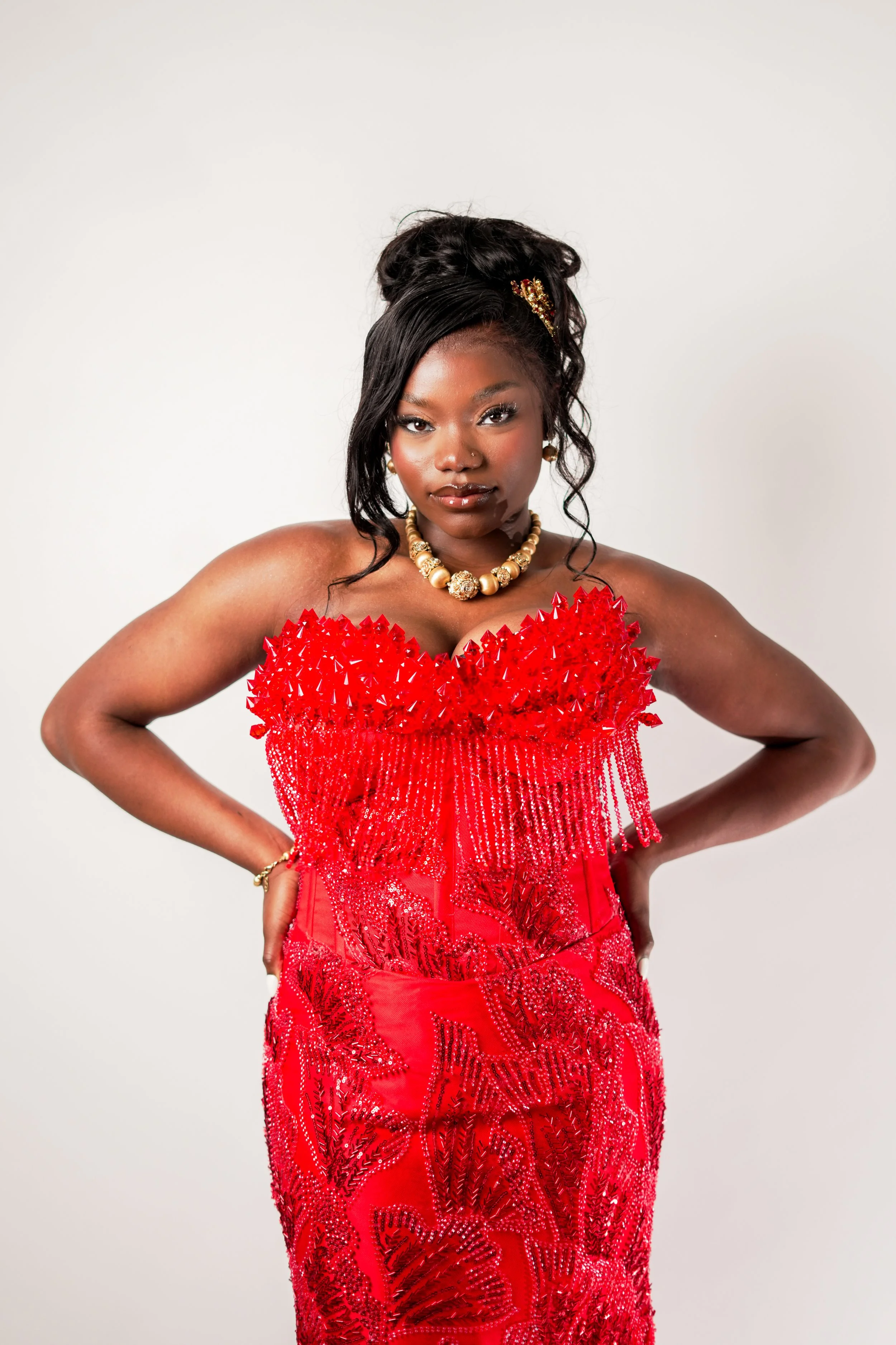 A woman with dark skin posing with her hands on her hips, wearing a red, embellished dress with a feathered neckline, pearl jewelry, and a decorative hairpiece, against a plain white background.