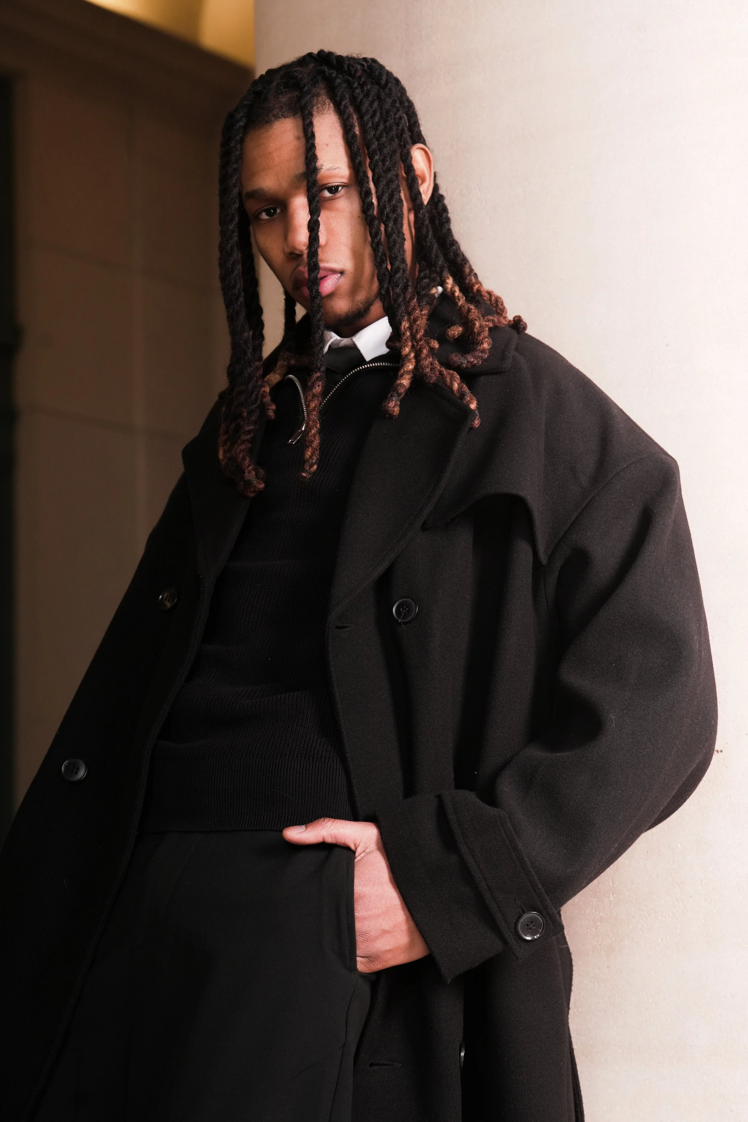 A young man with long dreadlocks standing against a white wall, wearing a black coat, white collared shirt, and black tie, with a serious expression. Editorial photography inspiration. Photoshoot. 