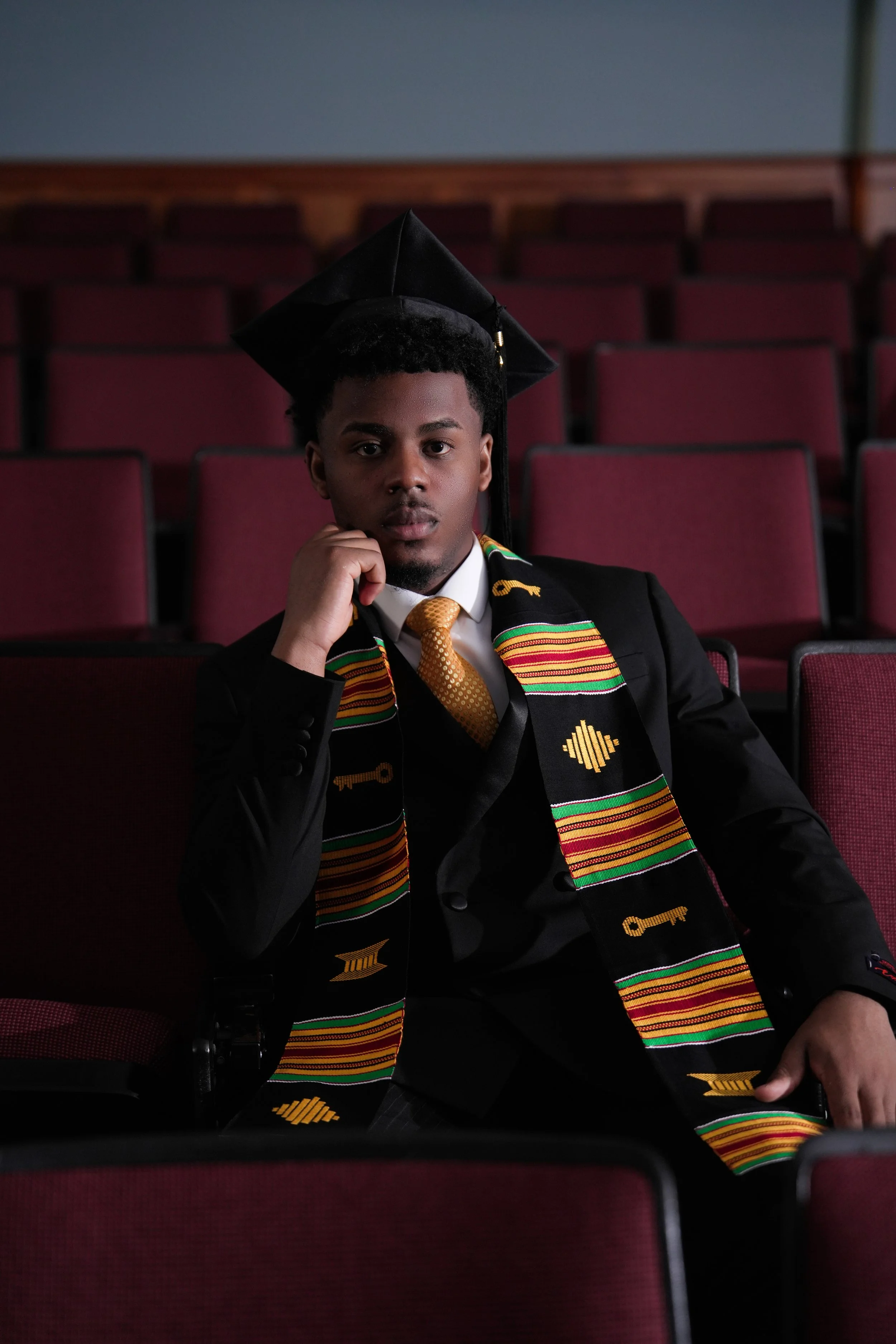 Young man in graduation cap and gown, sitting in an auditorium, wearing a colorful kente stole and gold tie, looking at the camera with a serious expression.