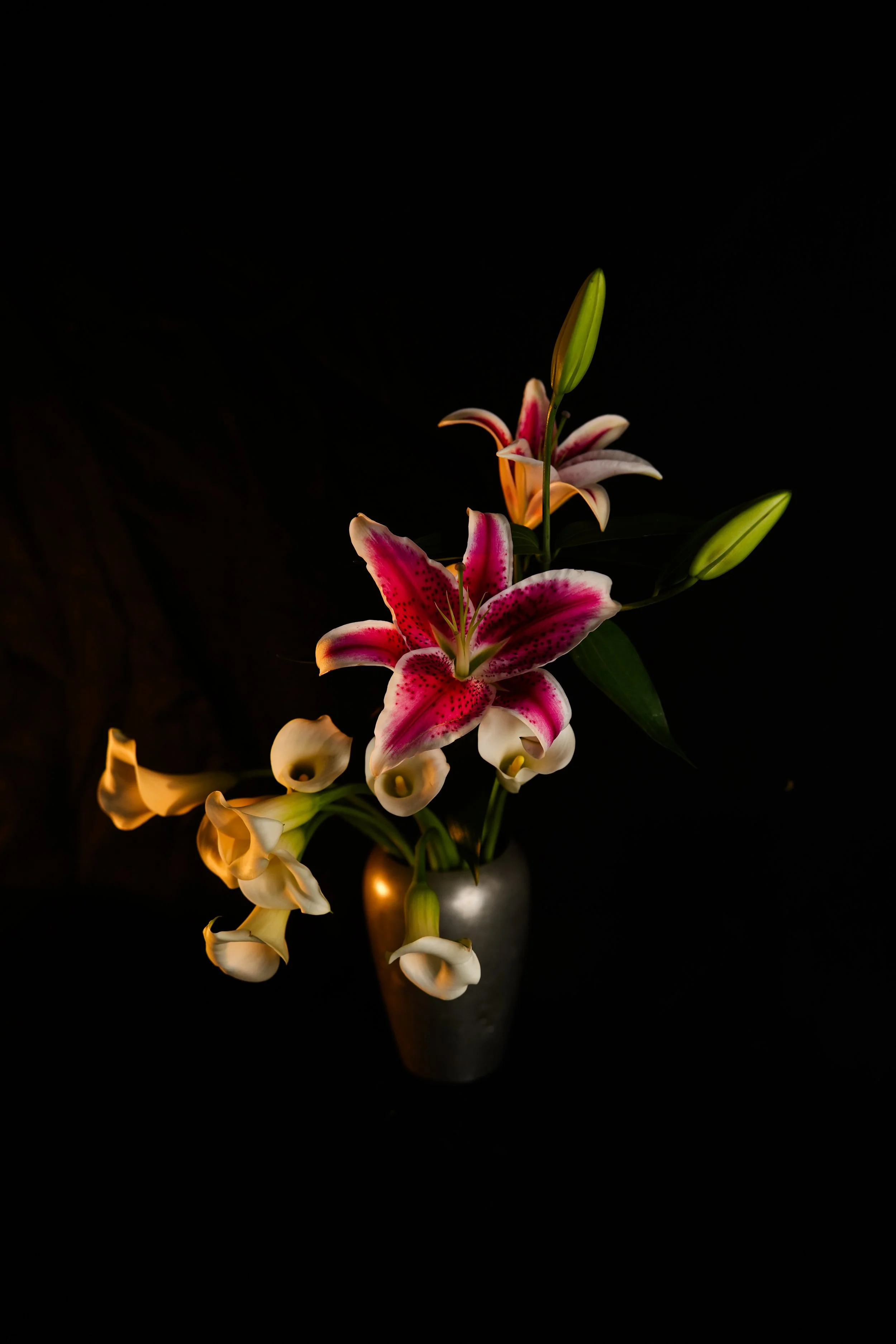 A bouquet of lilies featuring pink and white spotted lilies, orange lilies, and cream-colored calla lilies in a metallic vase with a dark background. product photography.
