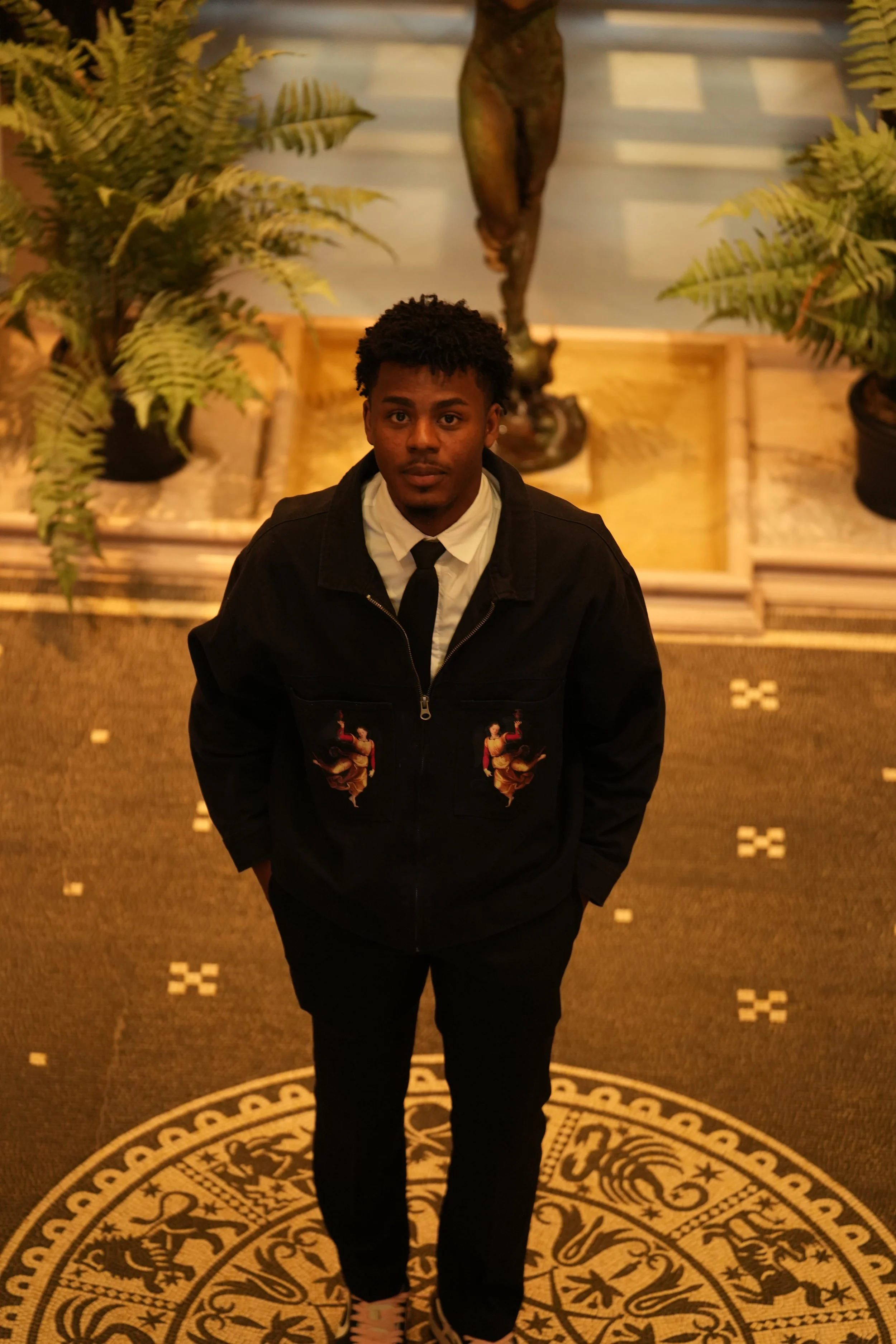 A young man with curly hair wearing a black jacket, white shirt, and black tie, standing on a patterned carpet in front of a backdrop featuring plants and a statue.