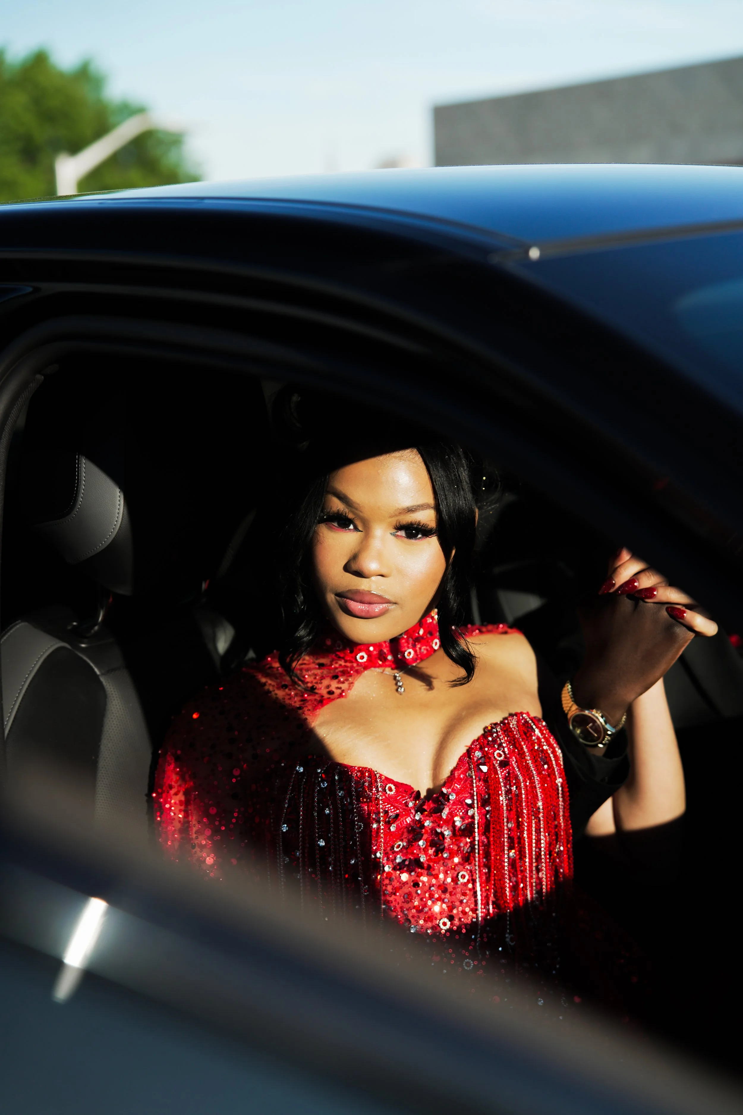 A woman in a red, sequined dress sitting inside a car, looking out the window, with her hand resting near her face. Prom Photography.