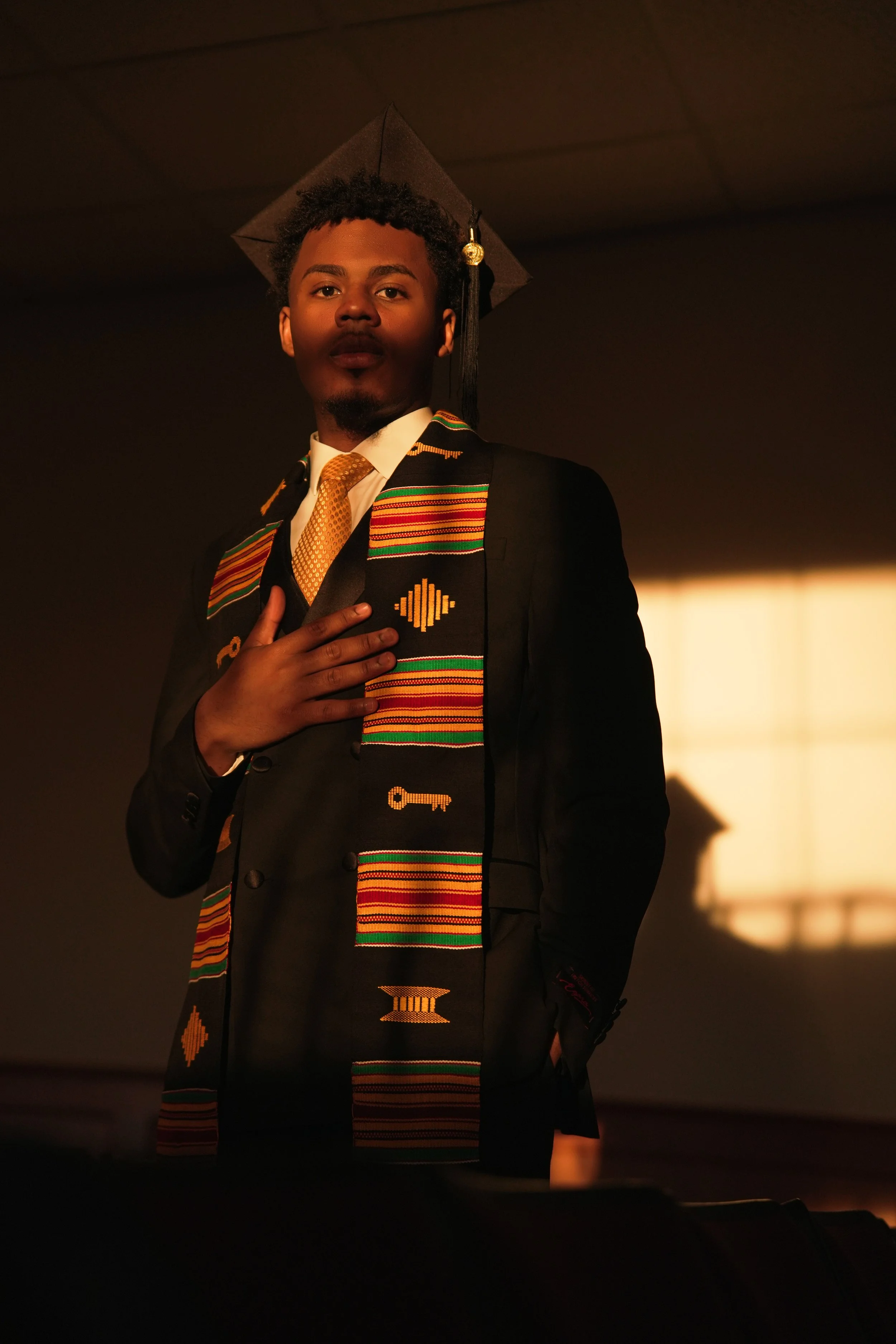 A young man in a graduation cap and gown with a colorful Kente cloth stole, standing with one hand over his heart, inside a dimly lit room.