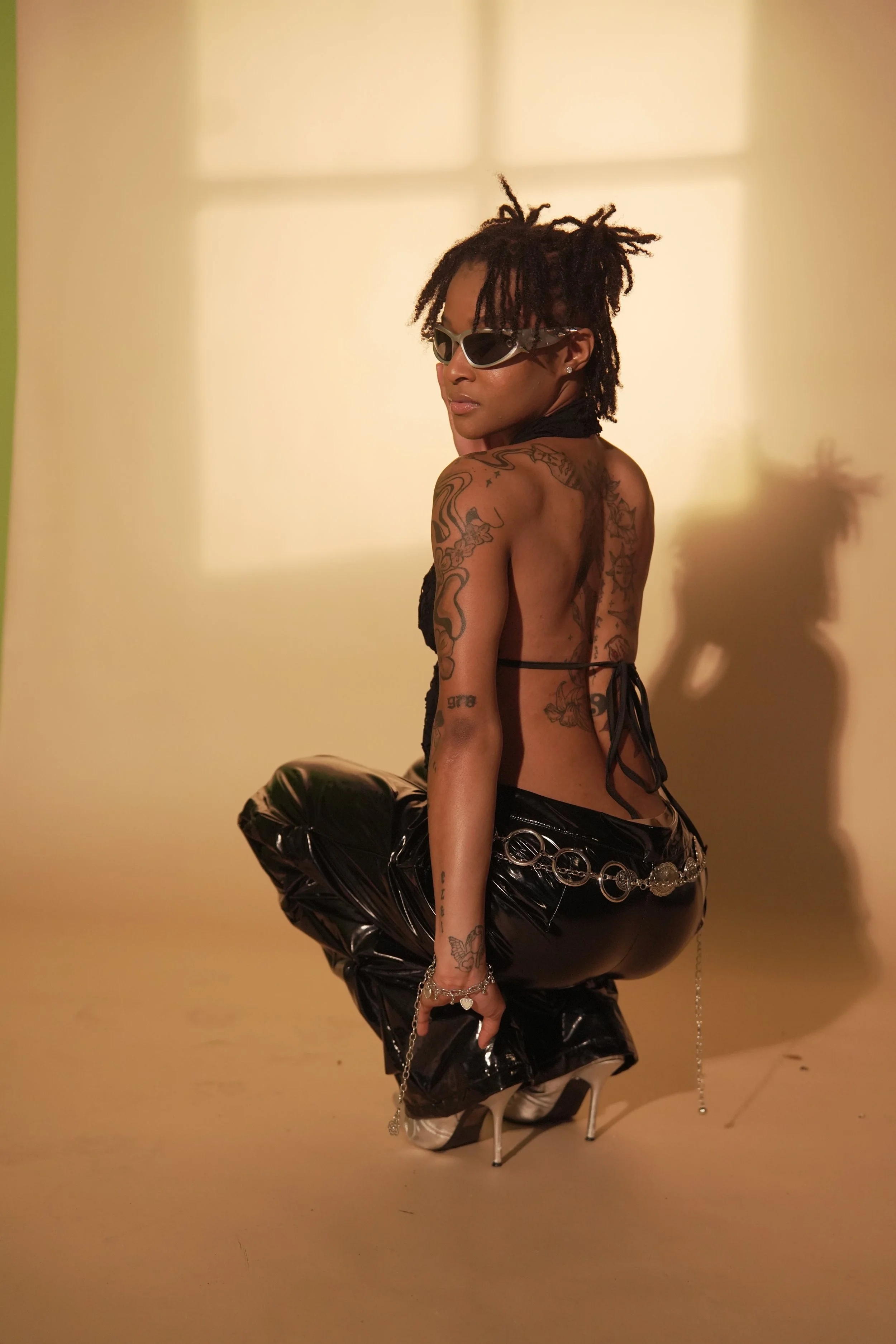 A woman with dreadlocks wearing sunglasses, a black top, shiny black vinyl pants, and high heels, striking a pose with one foot raised, in a studio with a plain yellow wall background.
