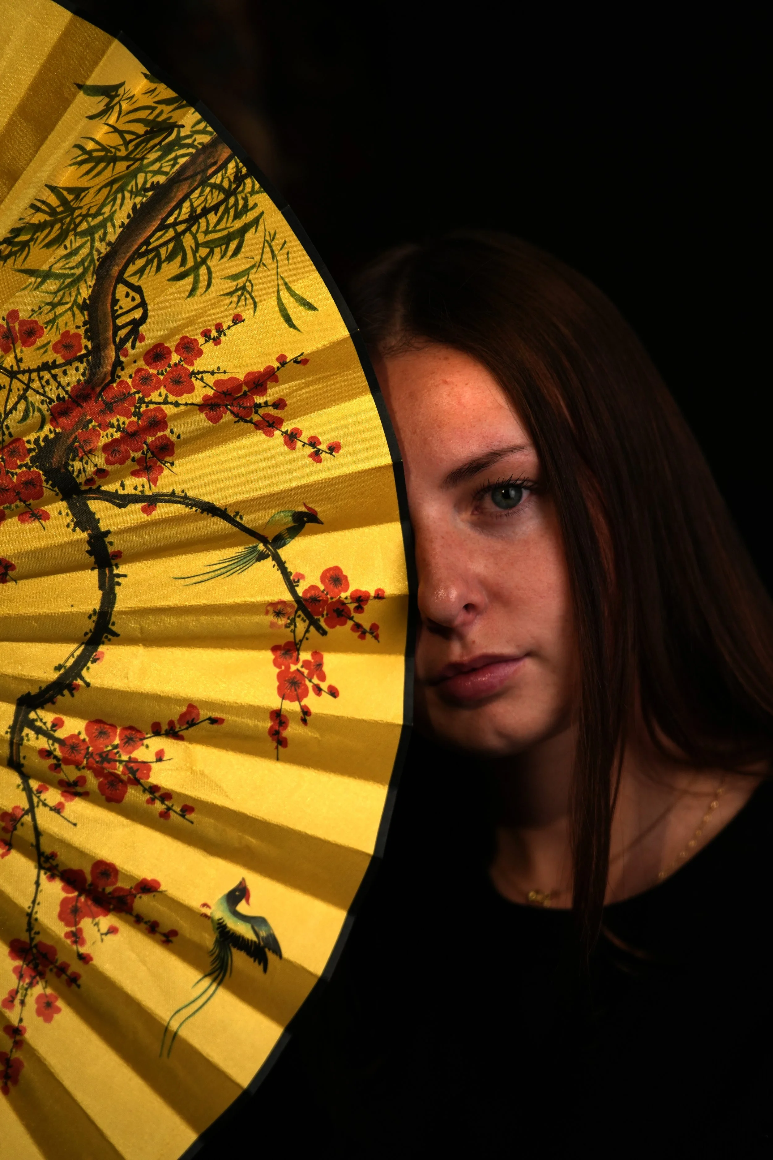 A woman with long brown hair and light skin holding a yellow fan decorated with red flowers and green branches, partially obscuring her face, with black background.