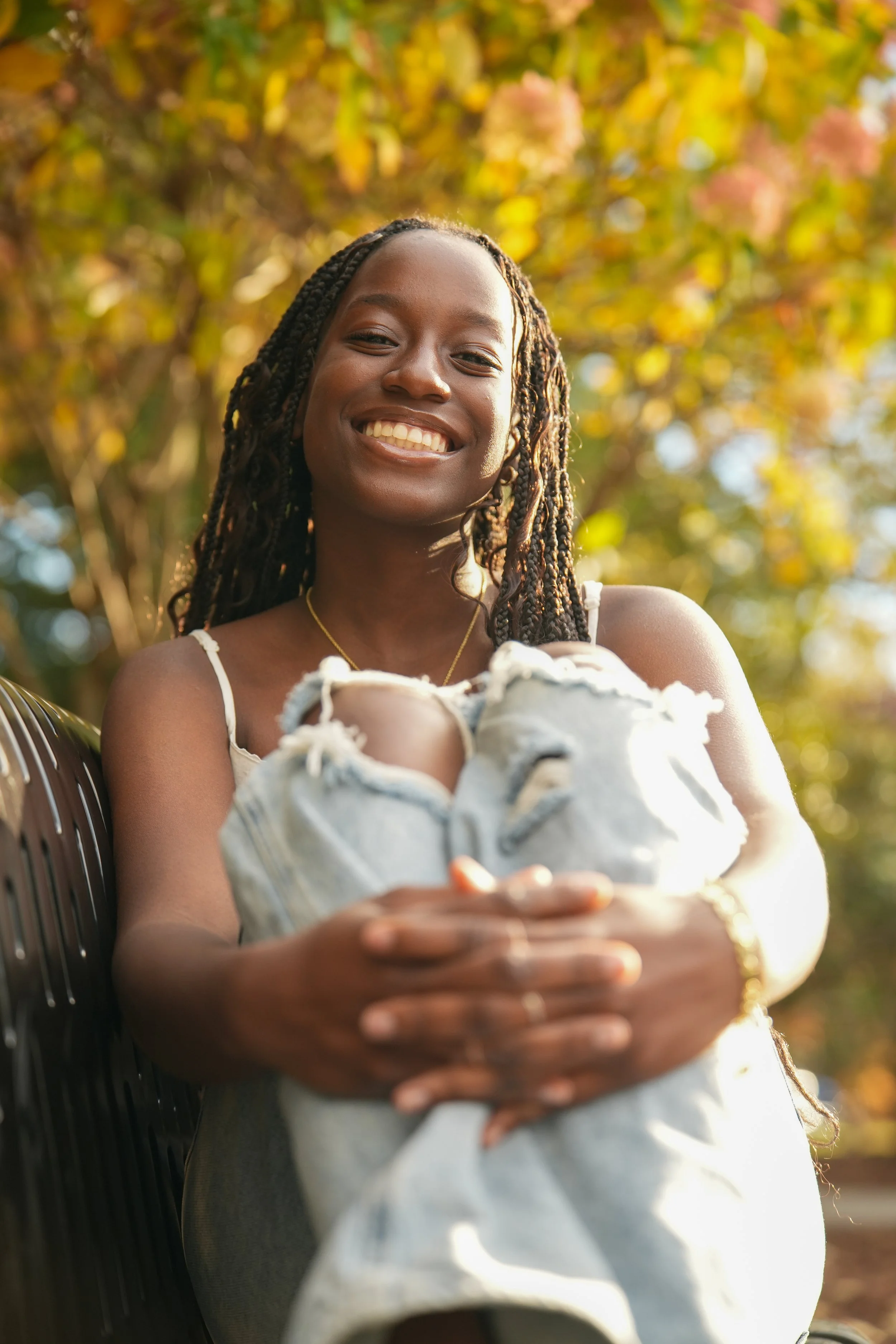 A woman smiling outdoors, holding a pair of worn denim shoes, with autumn leaves in the background. Senior Pictures 