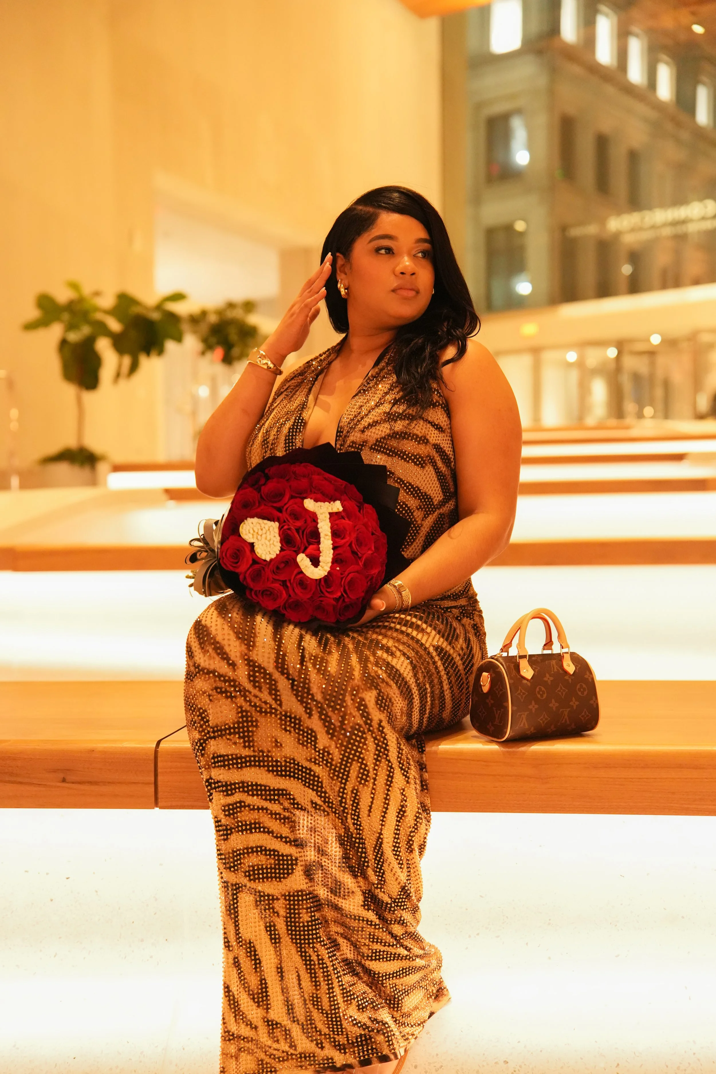 A woman dressed in a gold, animal print gown holding a bouquet of red roses with the letter 'J' on it, sitting on a bench indoors with modern decor and a large window in the background.