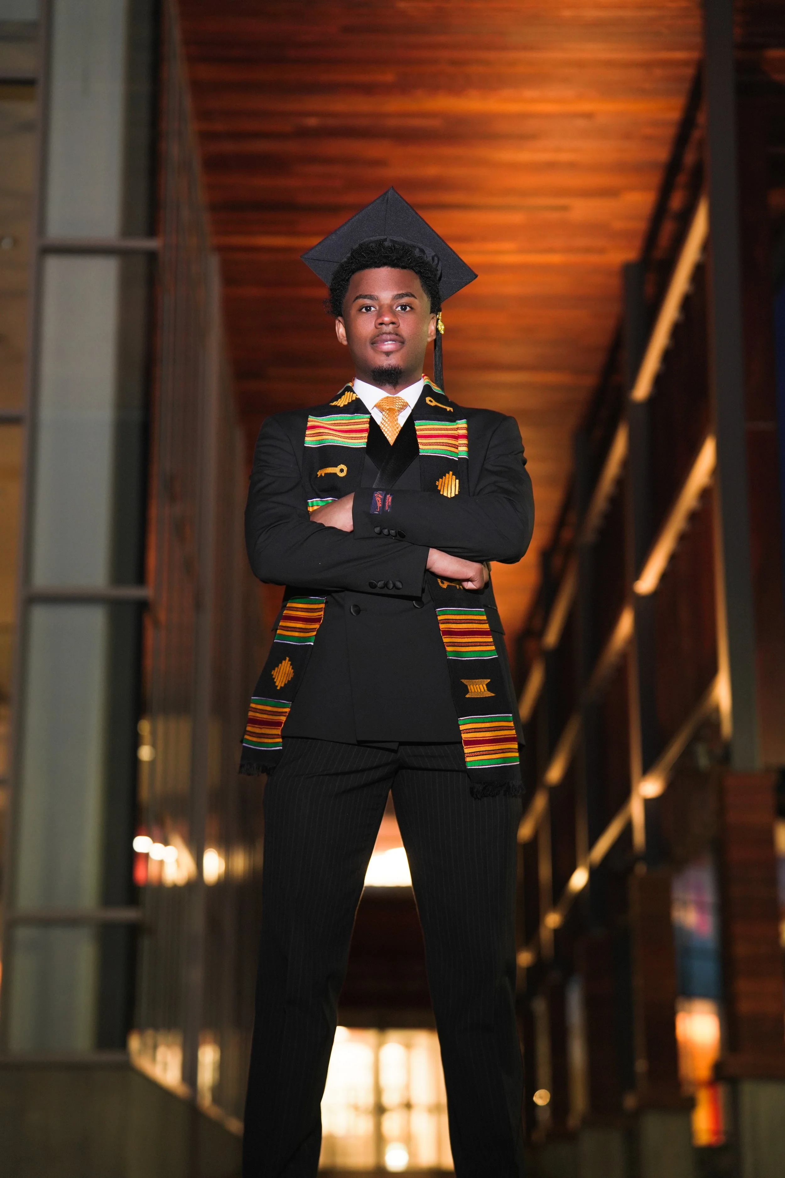 A young man in a graduation cap and gown with a colorful Kente stole, standing confidently with arms crossed inside a modern building with warm lighting and wooden ceilings. Graduation Pictures inspiration. Photography. photographer.