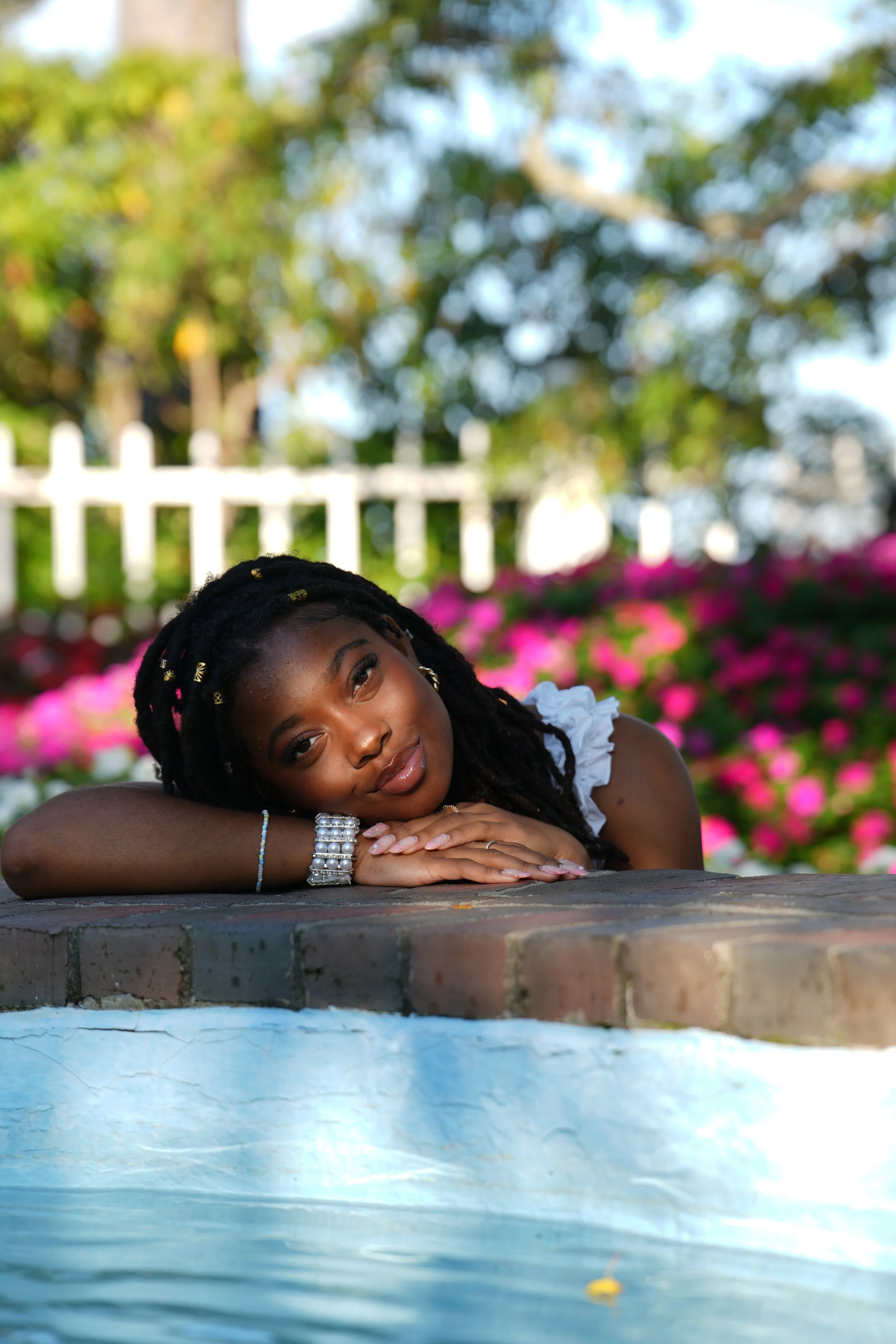 A young woman with braided hair and jewelry rests her head on her arms, lying on a brick ledge outdoors with pink flowers, trees, and a white picket fence in the background.