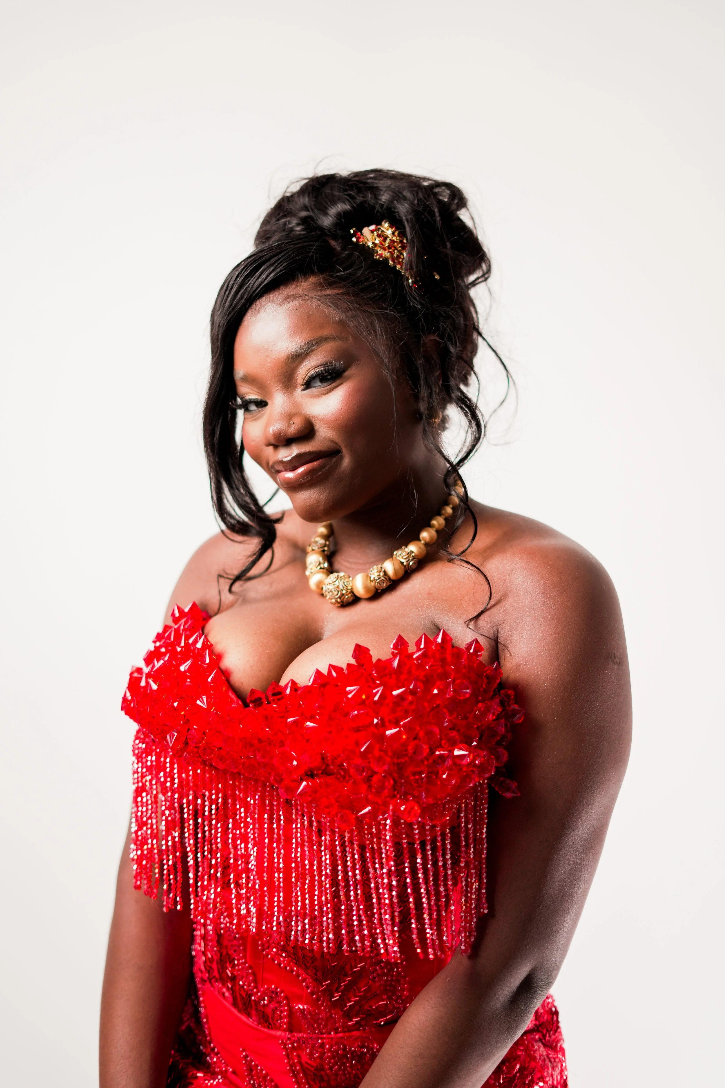 Portrait of a woman with dark hair styled with a decorative gold and red hairpin, wearing a bold red dress adorned with shiny beads and sequins, accessorized with a gold beaded necklace, posed against a plain white background.