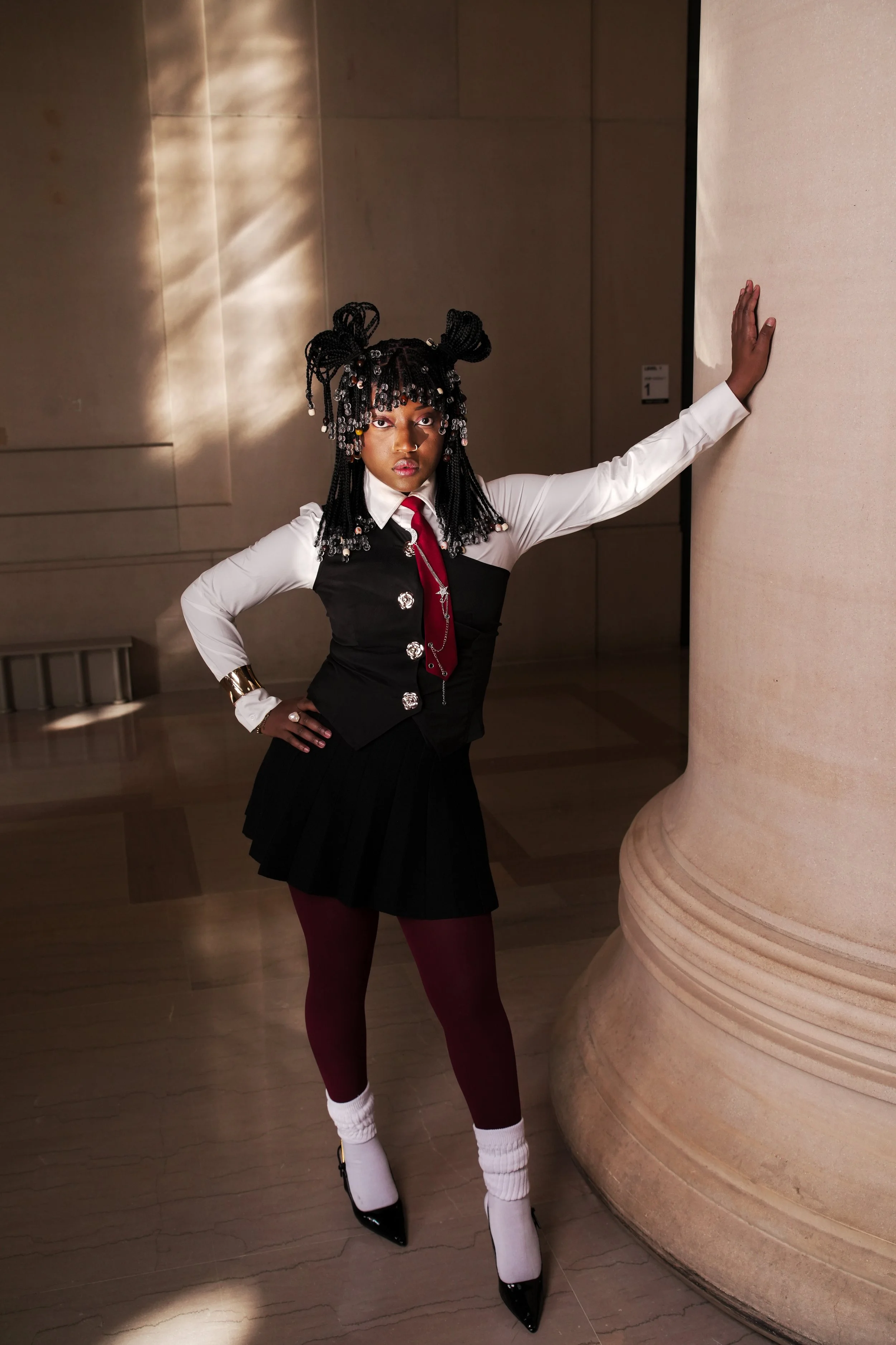 A woman in a fashionable outfit poses indoors, standing next to a large decorative column. She wears a white long-sleeve shirt, a black vest with silver buttons, a red tie, a pleated black skirt, maroon tights, white socks, and black high heels. Her 