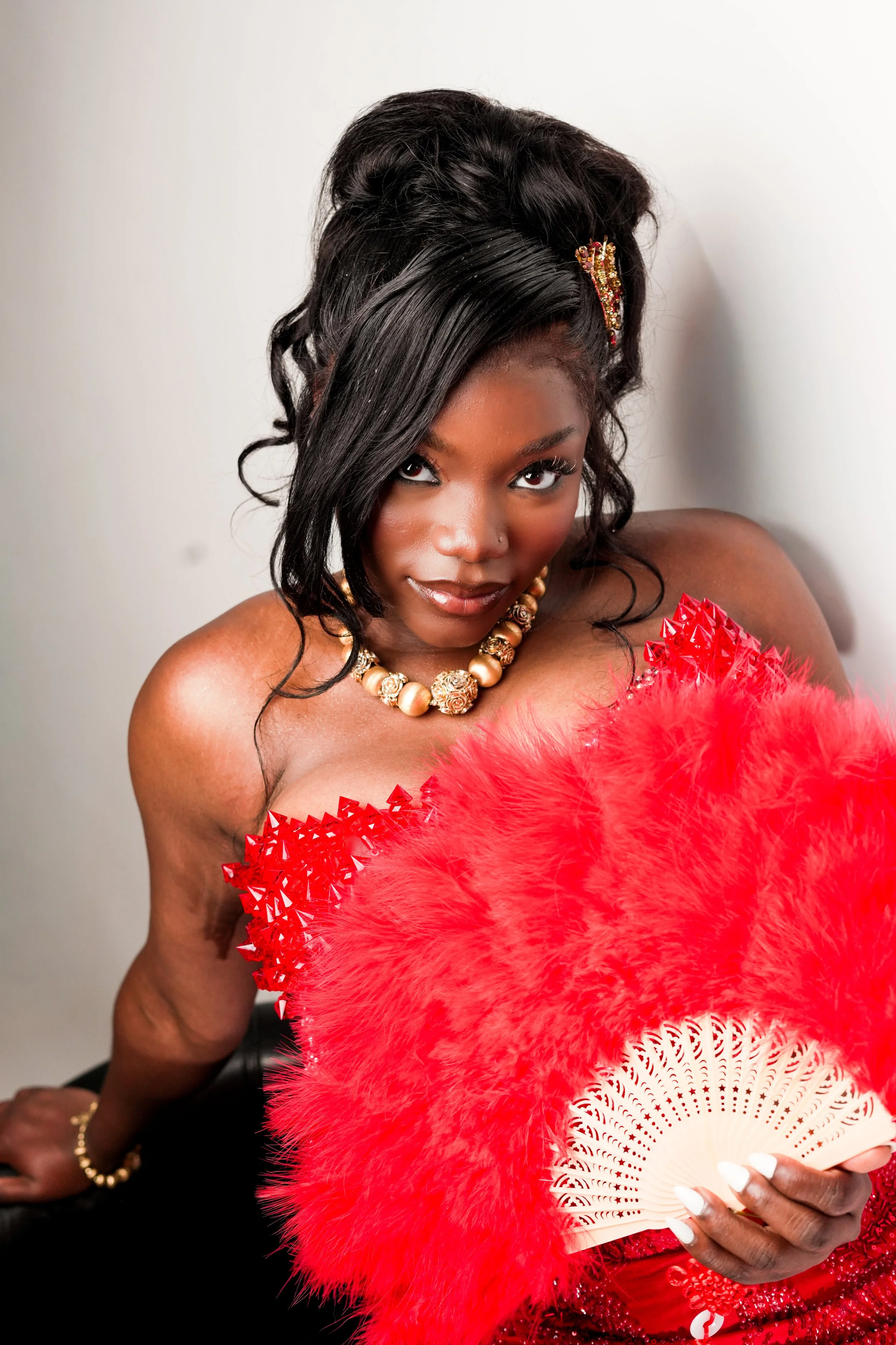 Portrait of an African American woman holding a cultural fan, wearing a red feathered garment, a pearl necklace, and a hair accessory, with a white background. Prom Photography. Birthday Photography