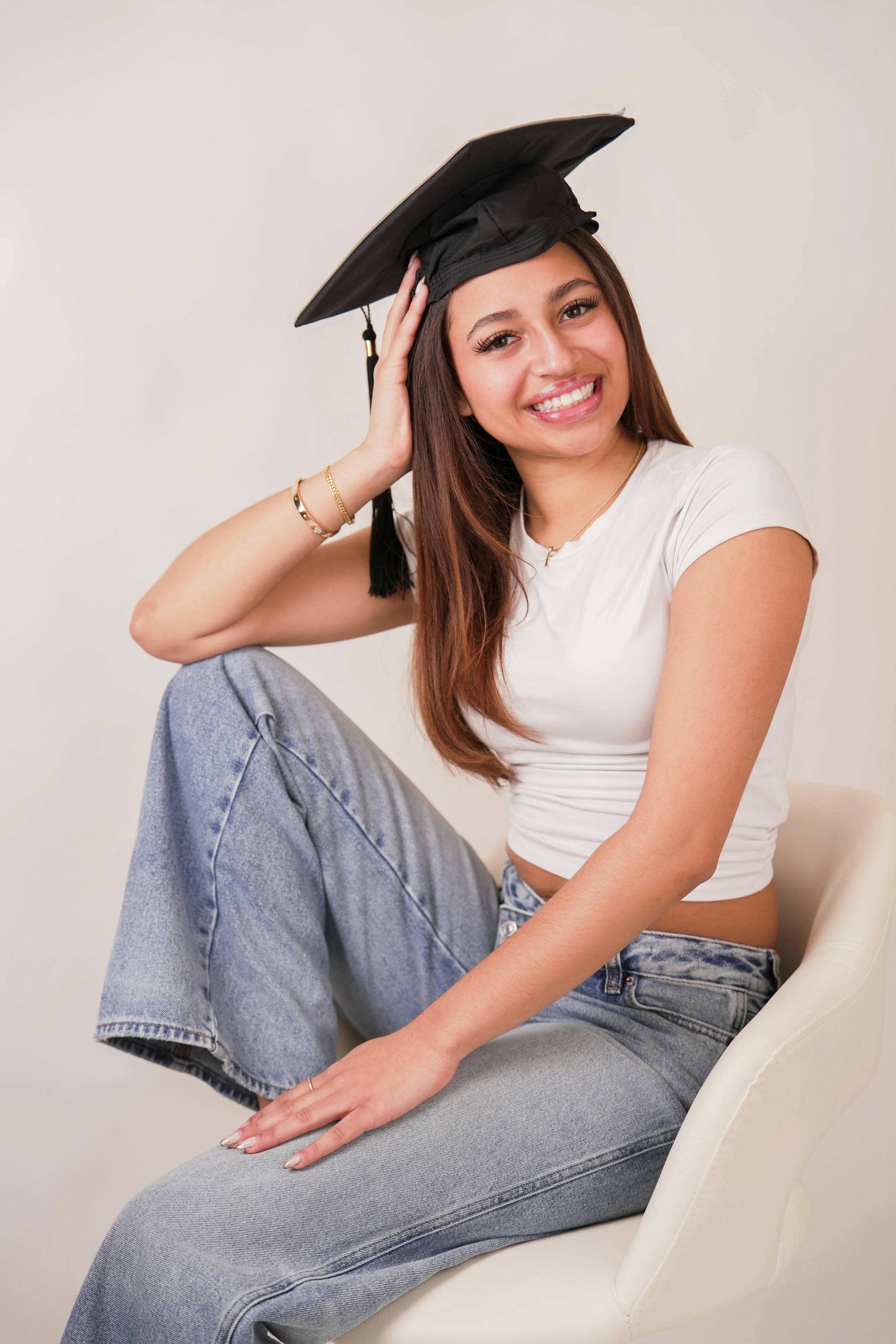 young woman with long brown hair, smiling, sitting on a white chair in front of a plain background. She is wearing a white crop top and light blue jeans, She has gold jewelry.  Best Graduation photoshoots.