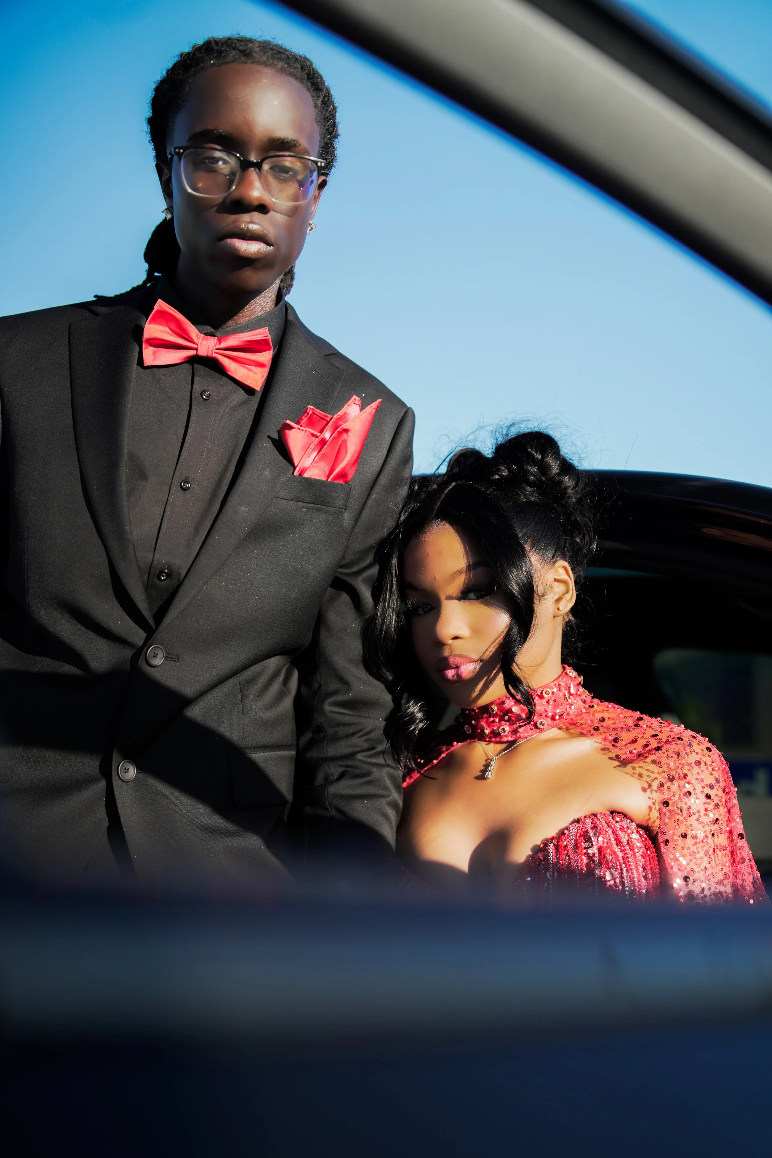 A man dressed in a black tuxedo with a red bow tie and red pocket square standing outdoors, partially obscured by a car window, next to a woman wearing a red sequined dress with an off-shoulder style, long dark hair, and makeup, with a clear blue sky