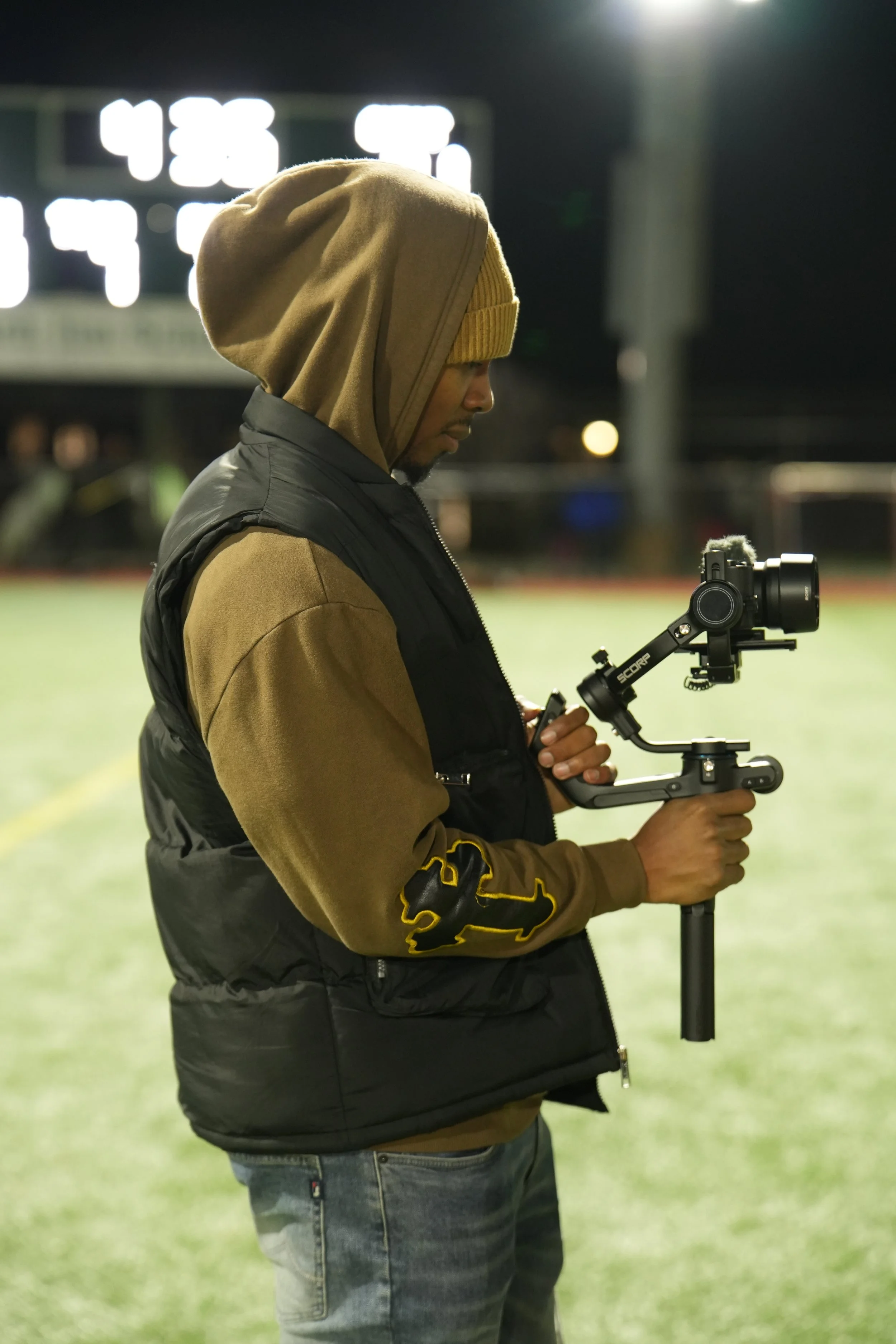 A man standing outdoors at night on a sports field, holding a camera stabilizer with a camera attached, wearing a brown hoodie under a black vest.