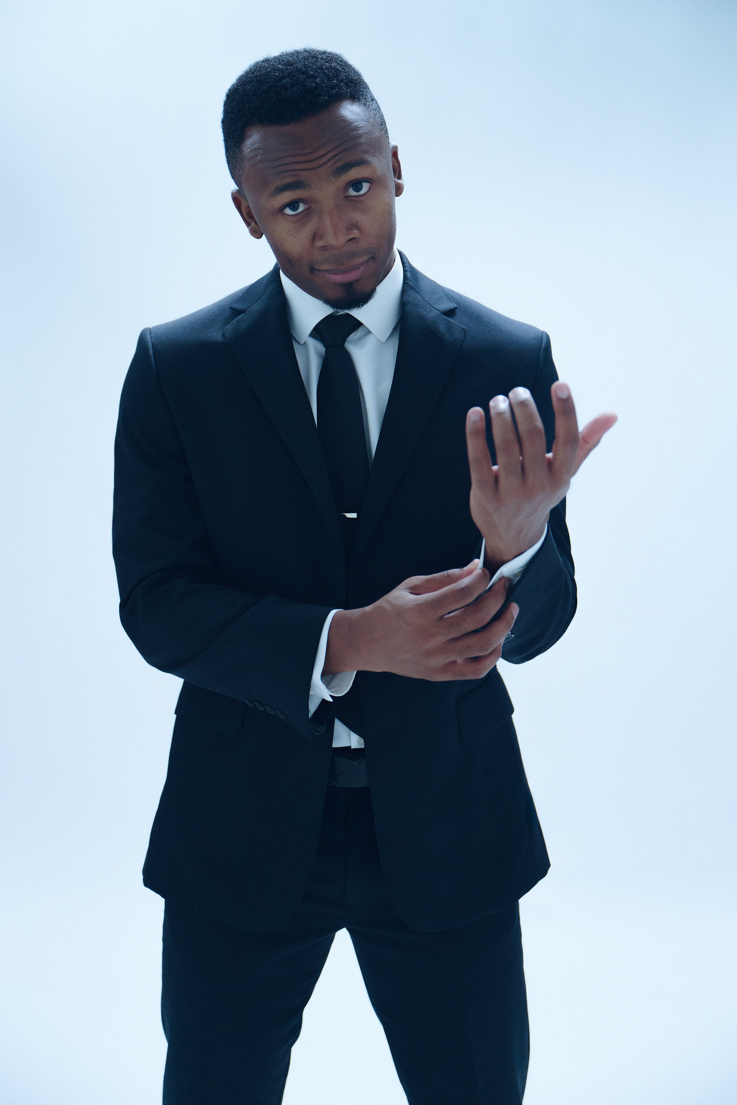 A young man with dark hair, wearing a black suit, white shirt, and black tie, standing against a plain light background. He has an expressive look, with one hand raised near his face.