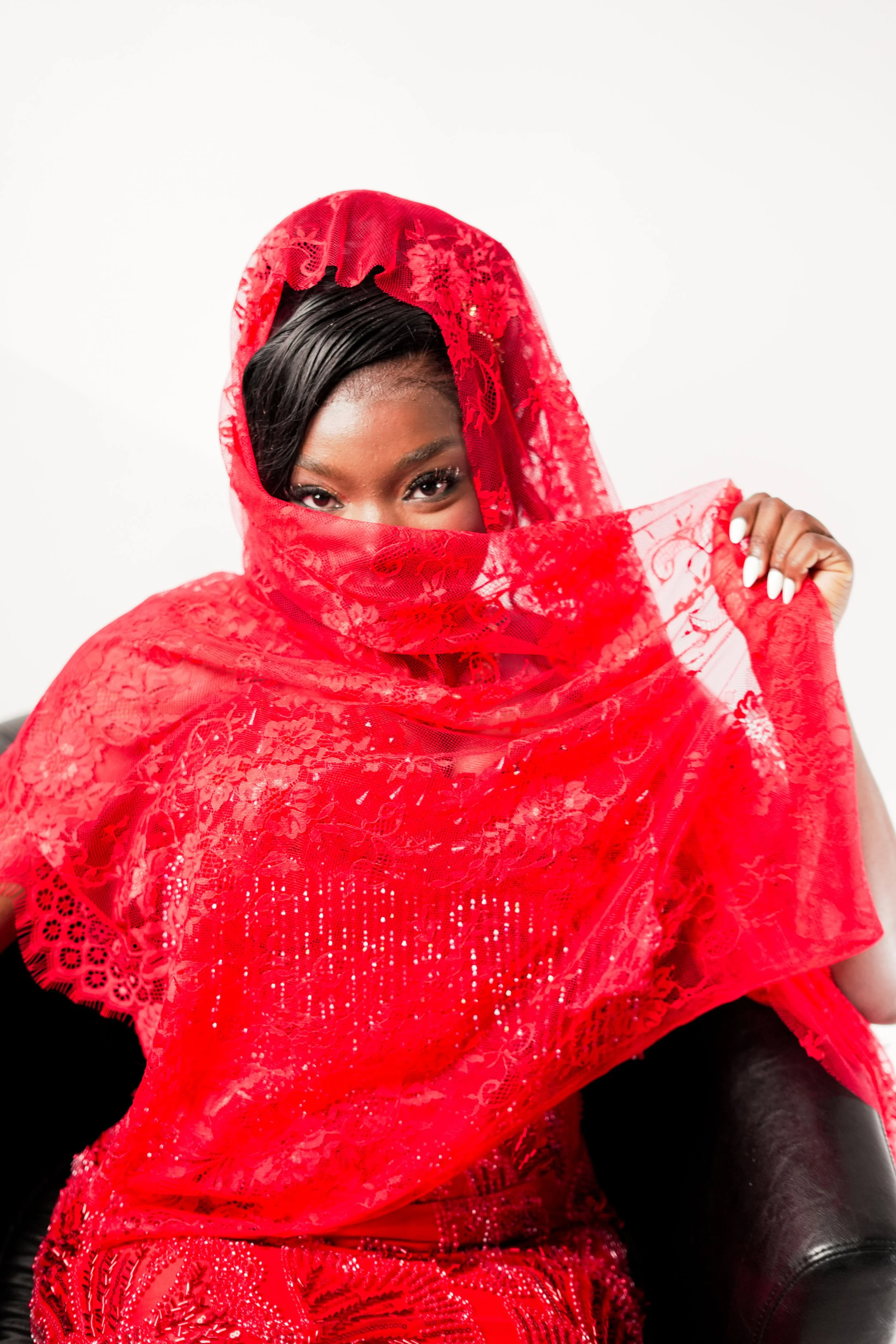 A woman with dark hair partially covered by a red lace shawl, looking directly at the camera with one eye visible, sitting on a black leather armchair.