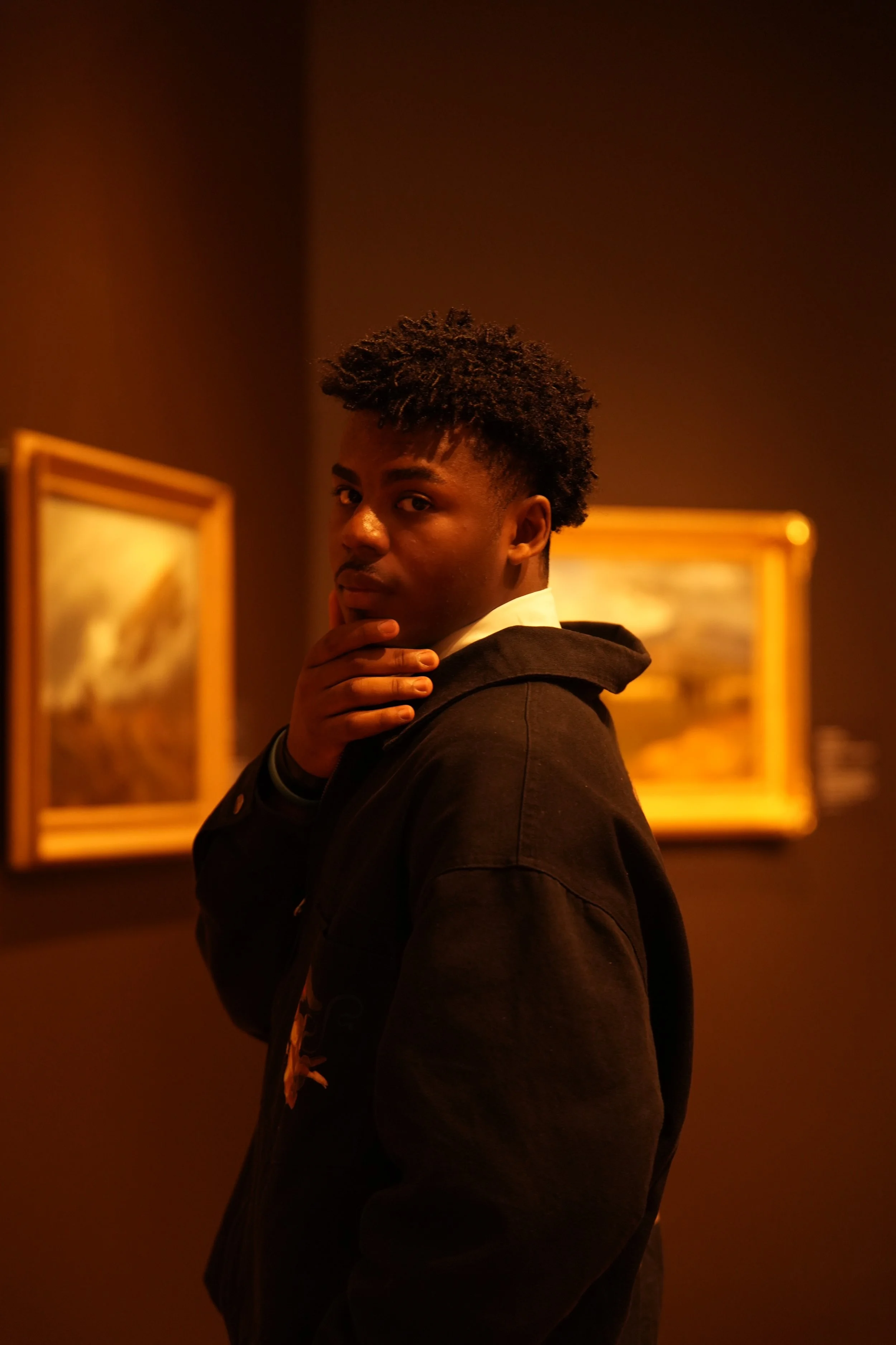 A young man with curly hair looking over his shoulder at the camera in an art gallery, with framed paintings on a dark wall behind him.