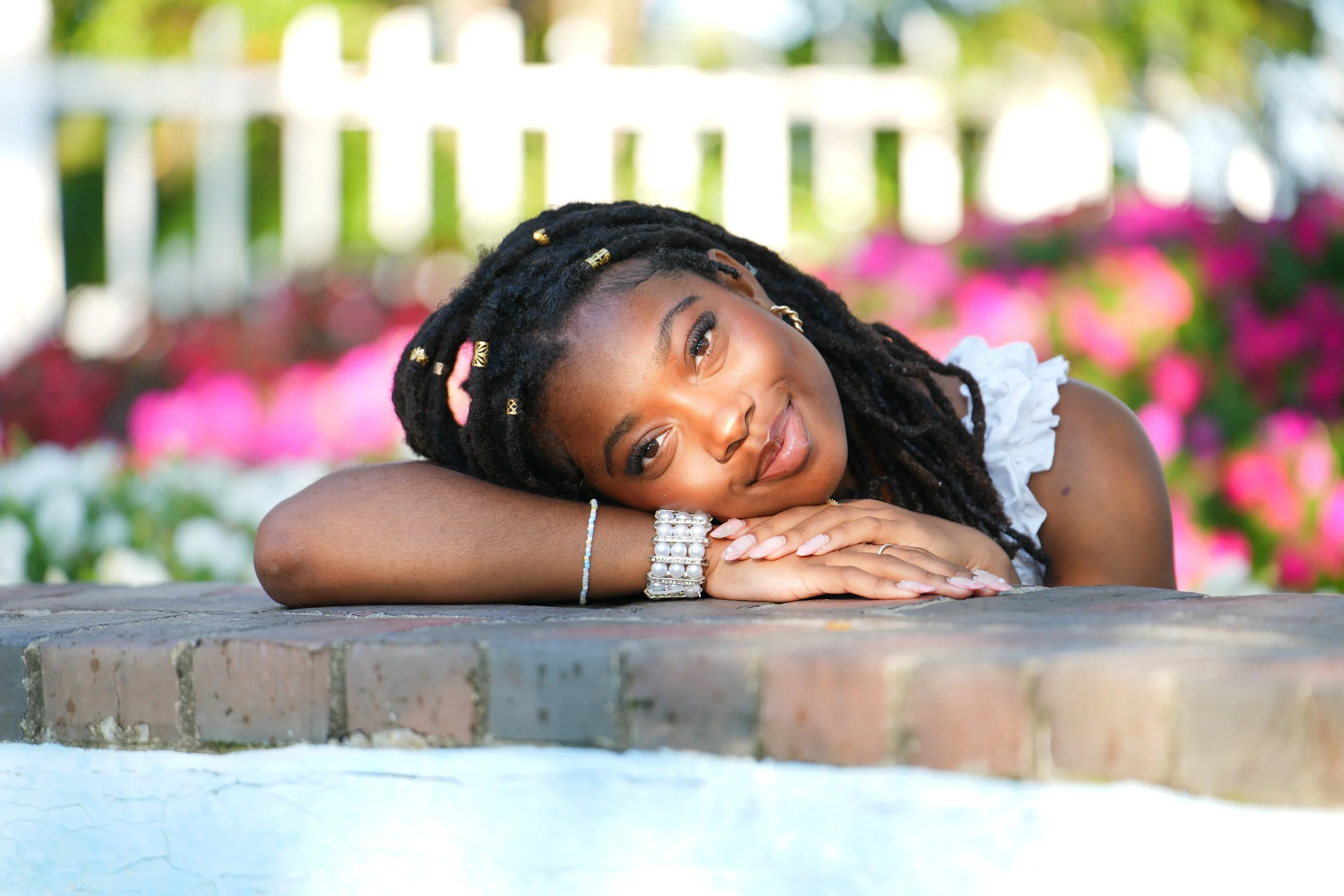 A woman with styled dreadlocks laying on a brick surface outdoors, smiling softly with her head resting on her folded arms, surrounded by colorful flowers.