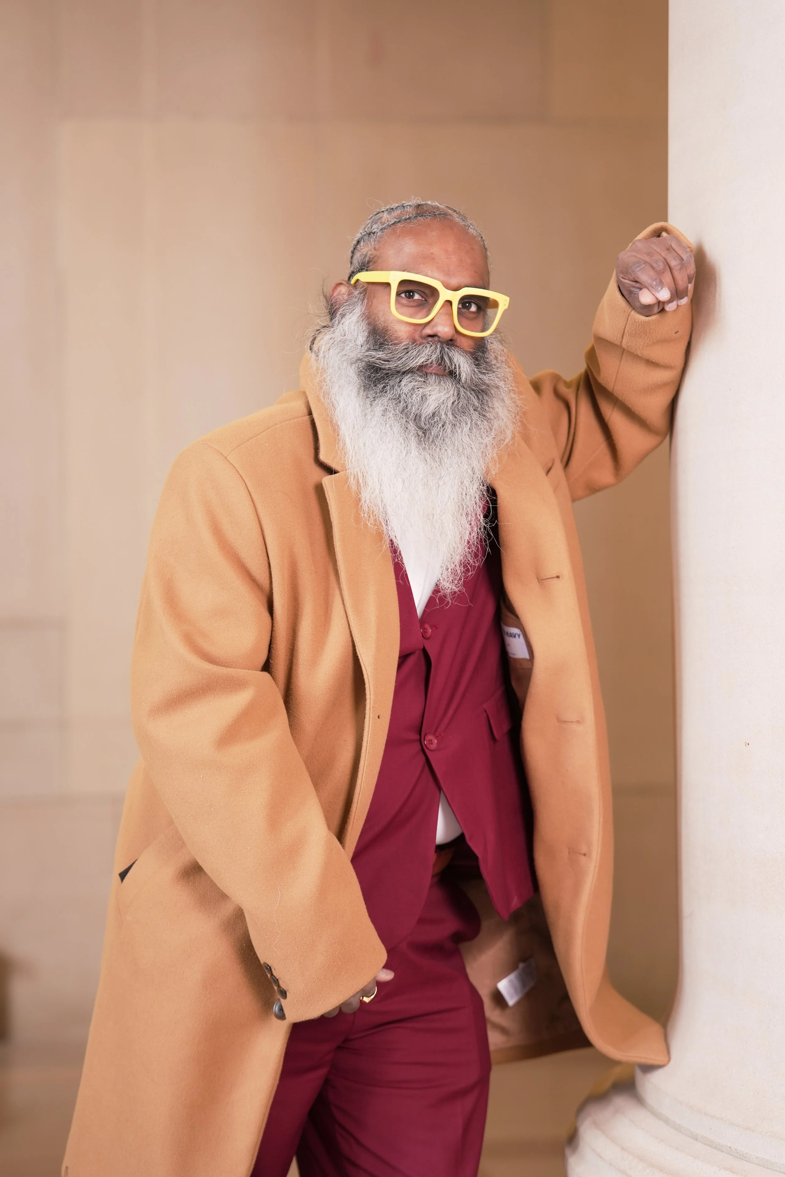 A man with a long gray beard, gray hair, and wearing yellow glasses, a beige coat, and a maroon shirt, leaning against a white column in an indoor setting.
