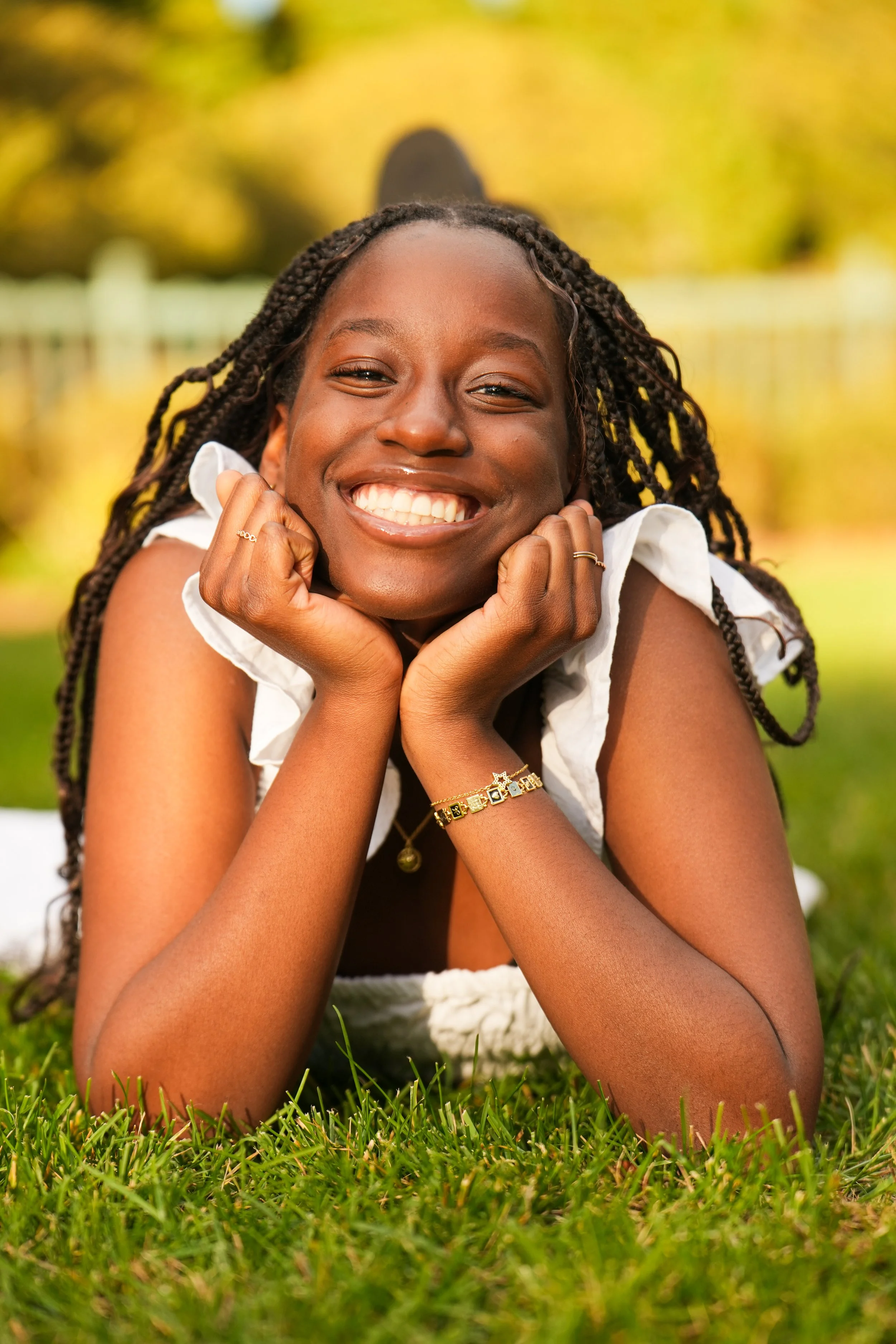 A young woman with braids smiling and resting her chin on her hands, lying on green grass in an outdoor setting with blurred trees and yellow foliage in the background.