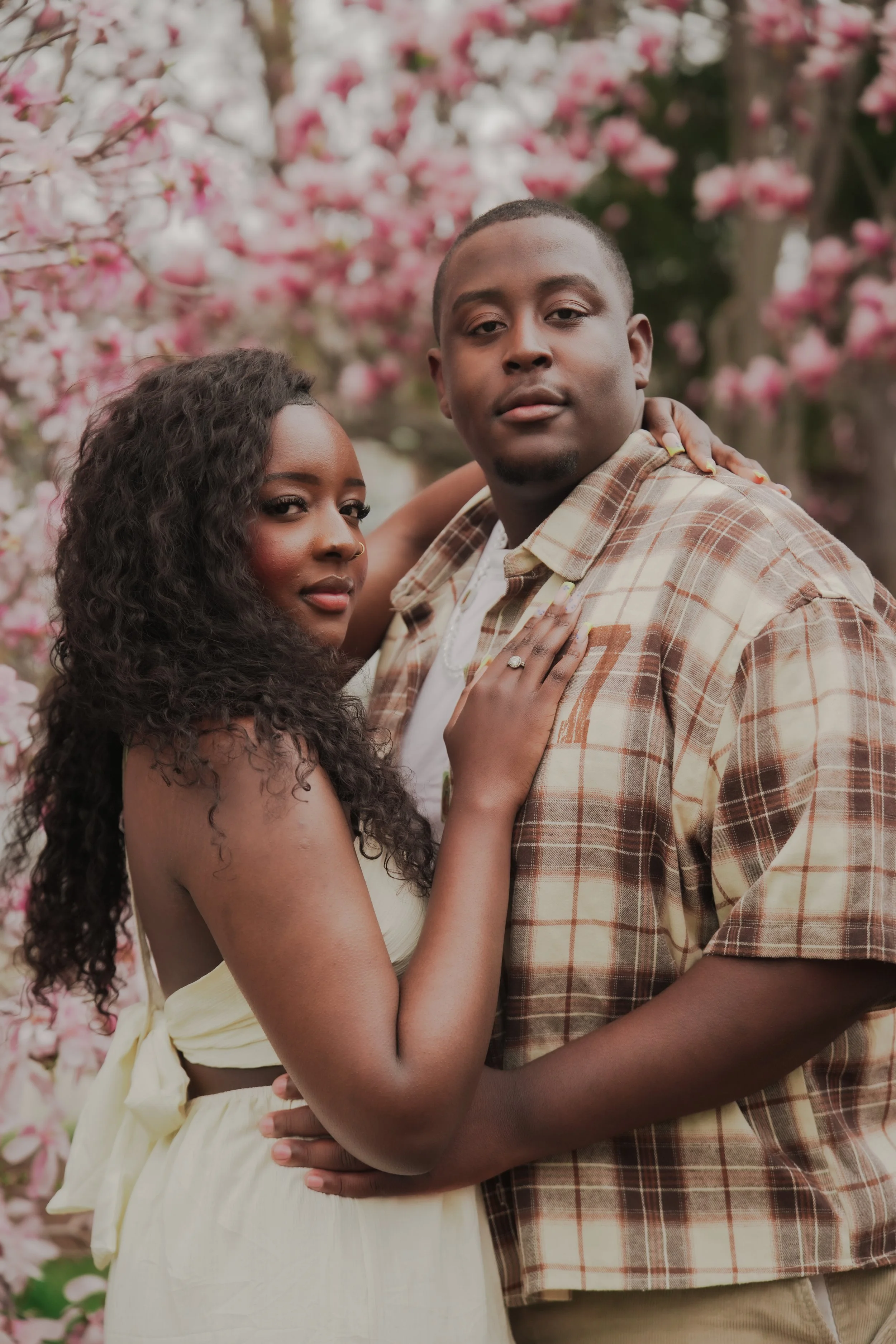 A young Black man and woman standing close together in front of pink blossoming trees. The woman has curly hair, and has her hand on the man's chest. gently holds the woman around her waist. sharing an intimate moment. Best Couples photoshoot inspo.