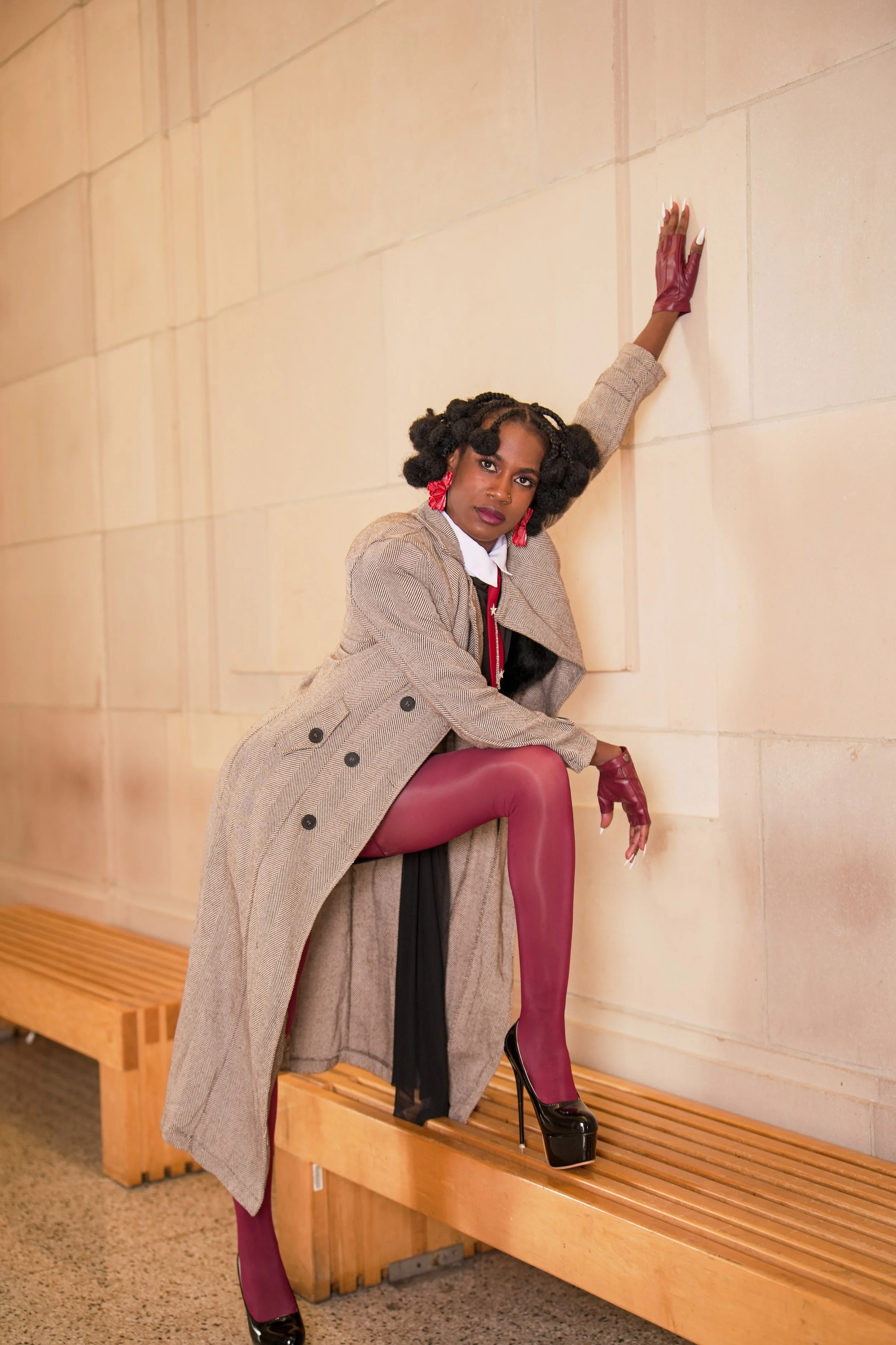 A woman with natural hair styled in bantu knots, wearing a beige plaid coat, white shirt, red lipstick, red earrings, and matching red gloves. She is posing against a beige wall, with one arm extended upward and the other resting on a bench. She has 