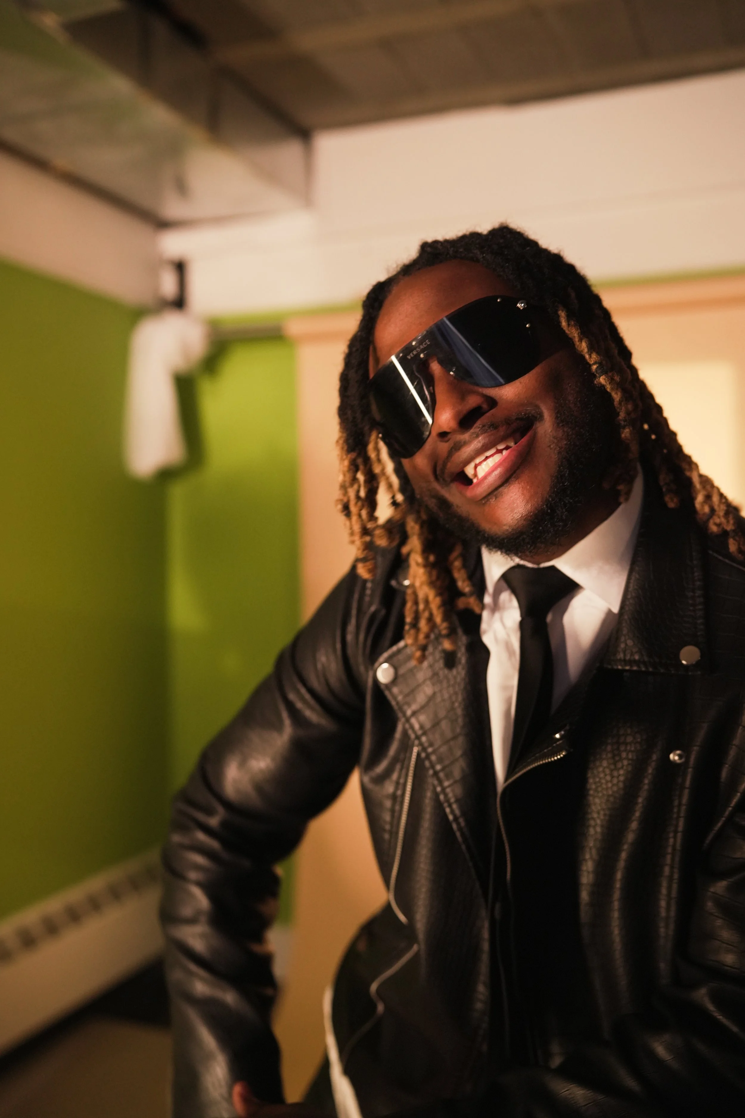 A man with dreadlocks wearing sunglasses and a black leather jacket, smiling, leaning slightly forward in a colorful room with a green wall and a white curtain.