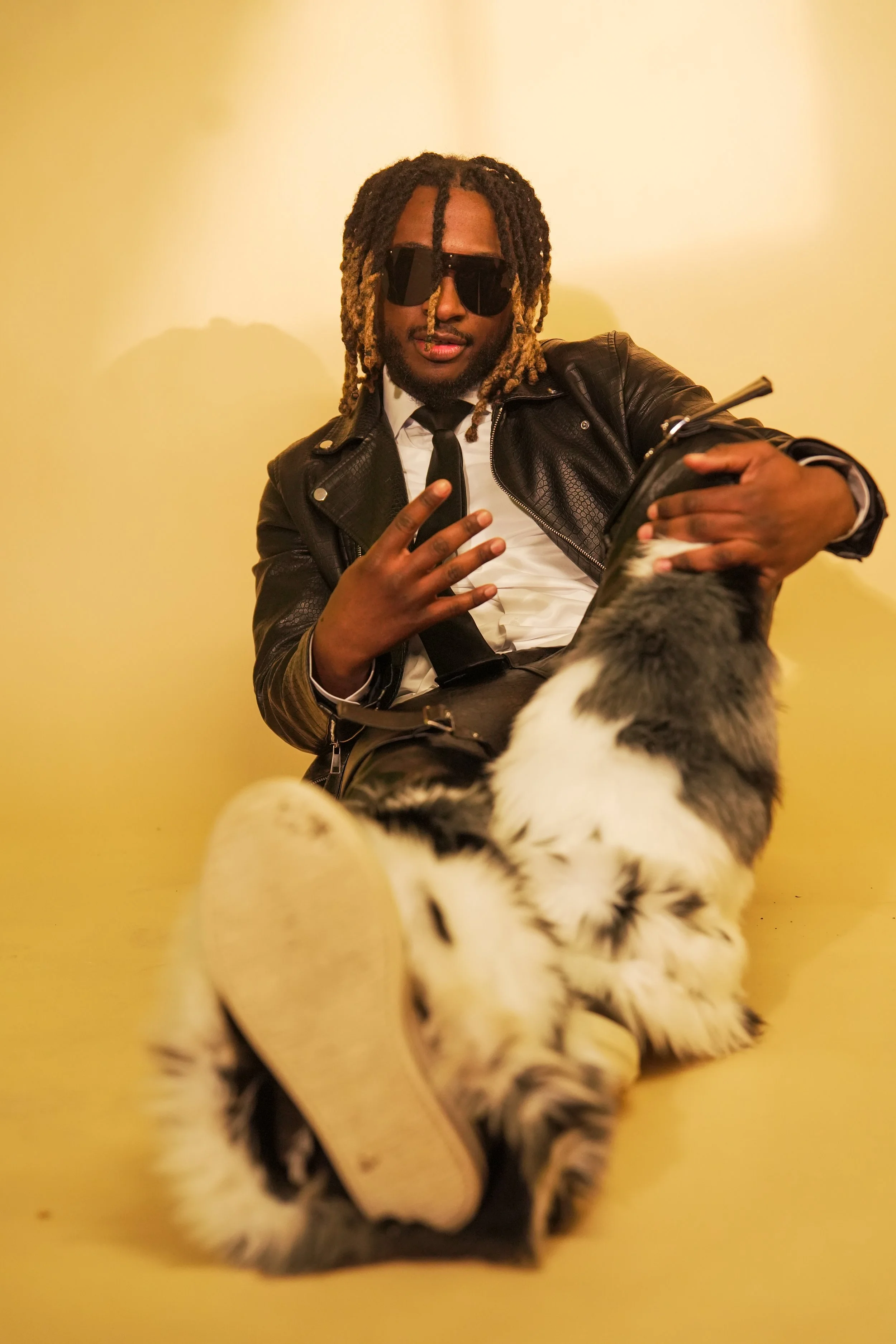 A man with dreadlocks, wearing sunglasses, a white shirt, a black leather jacket, and a tie, sitting against a yellow wall, with a dog in the foreground.